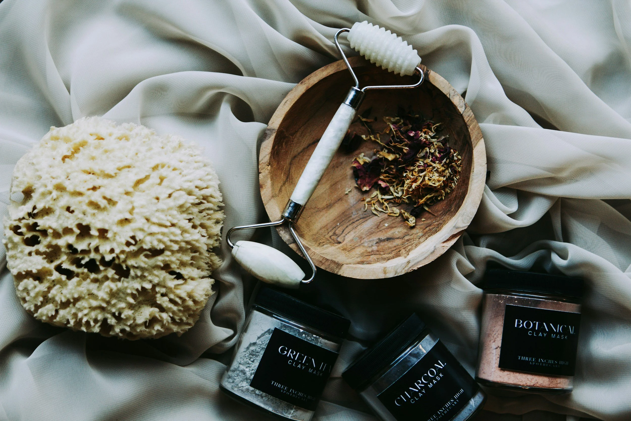 Various skincare products including charcoal and green tea clay masks, a natural sponge, and a sponge roller on a soft, light-colored fabric background.