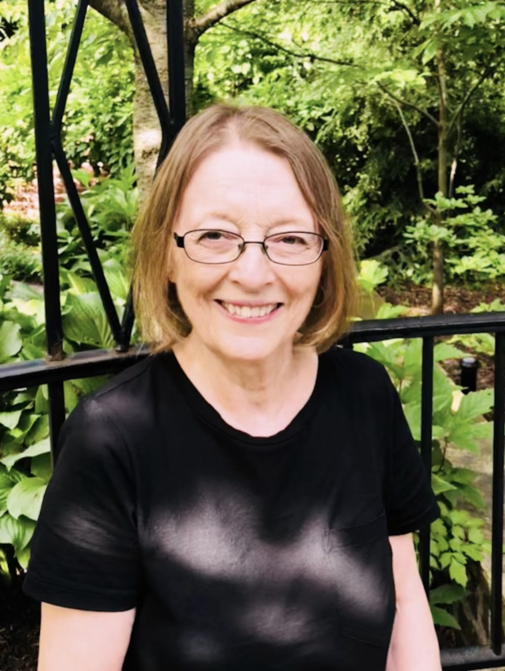 A smiling woman with glasses and shoulder-length hair, wearing a black t-shirt, outdoors in a garden with green foliage and trees in the background.