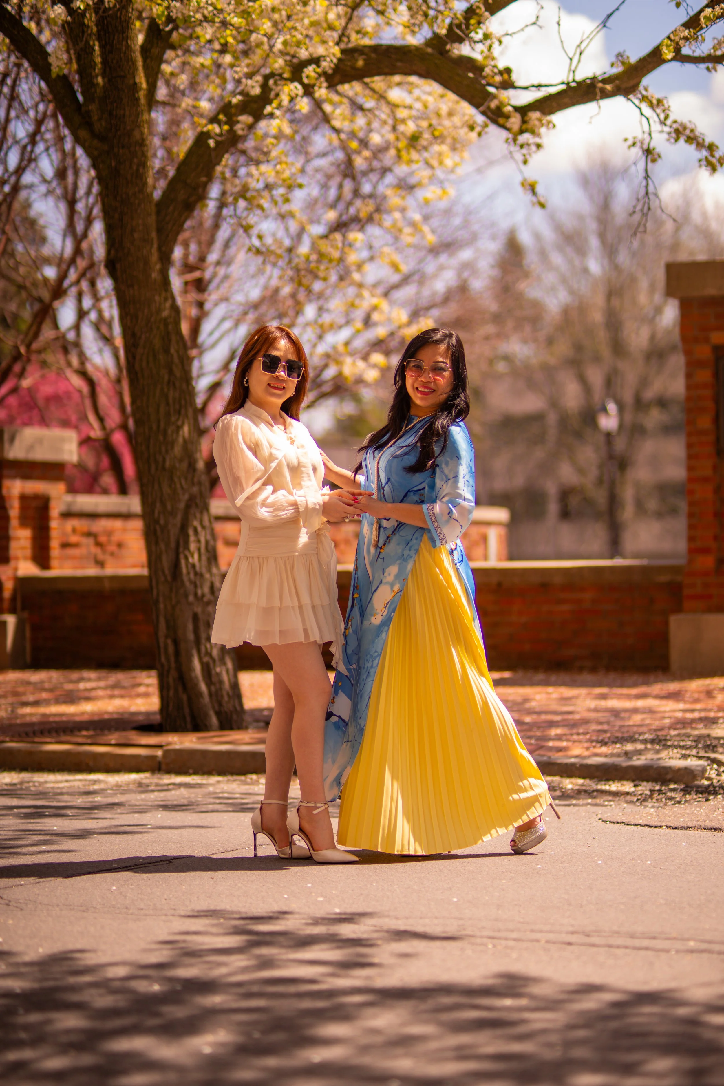 Two women standing together under a tree with blooming flowers, smiling and holding hands. One woman is wearing a white dress and high heels, the other is wearing a blue and yellow dress with a pleated yellow skirt.