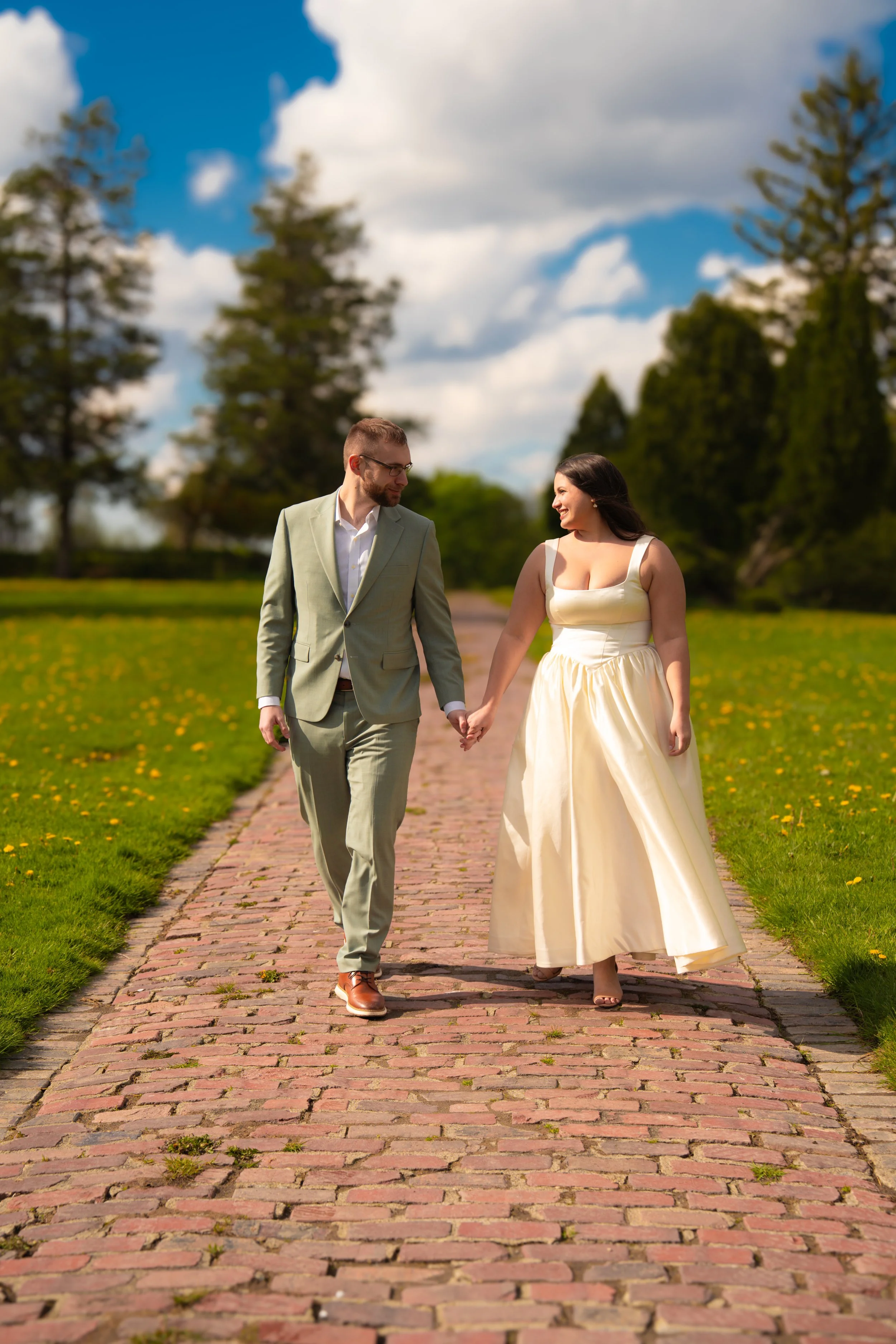 A couple dressed in formal attire, walking hand-in-hand on a cobblestone path in a park with green grass and tall trees under a partly cloudy sky.