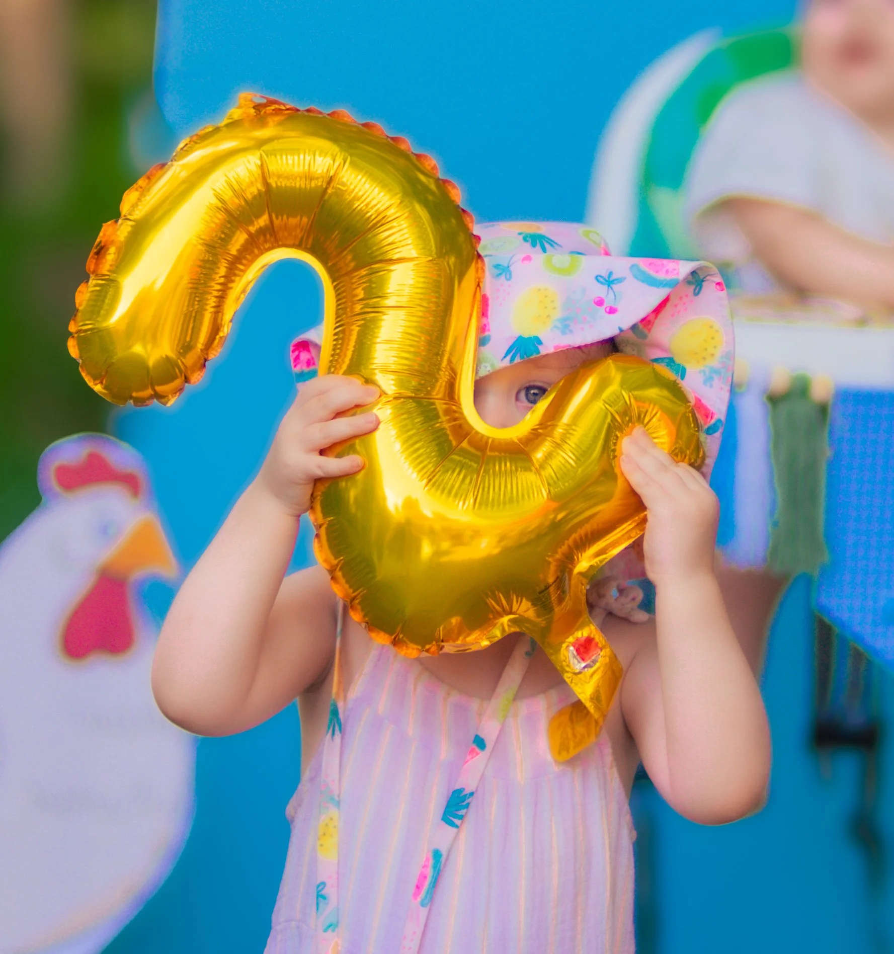 Child holding a gold foil number 0 balloon, wearing a colorful dress with yellow and blue pattern and a pink hat with blue flowers, at a birthday celebration.