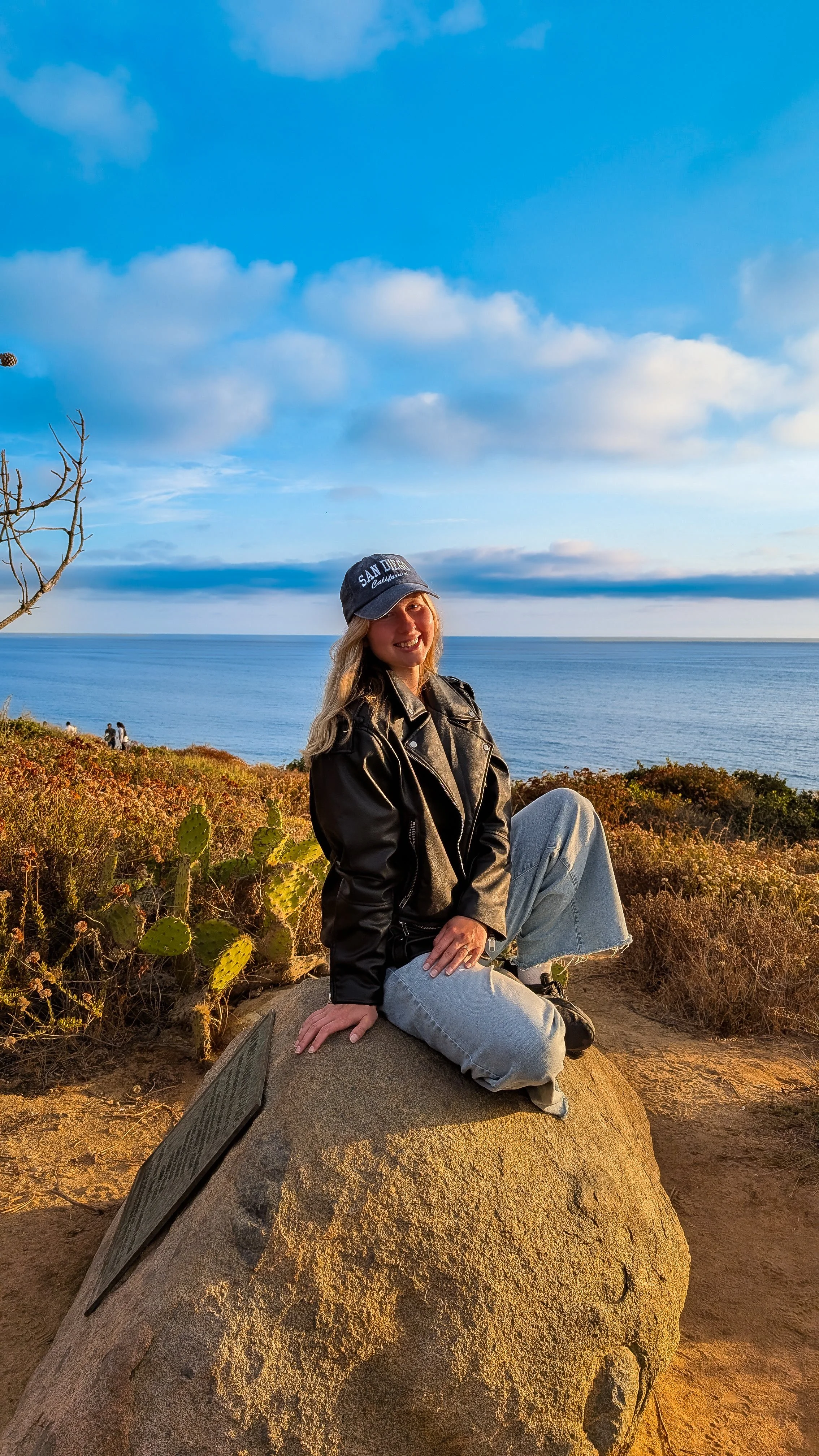A woman sitting on a large rock in a natural landscape with oak and cactus plants, overlooking the ocean under a blue sky with clouds, during sunset.