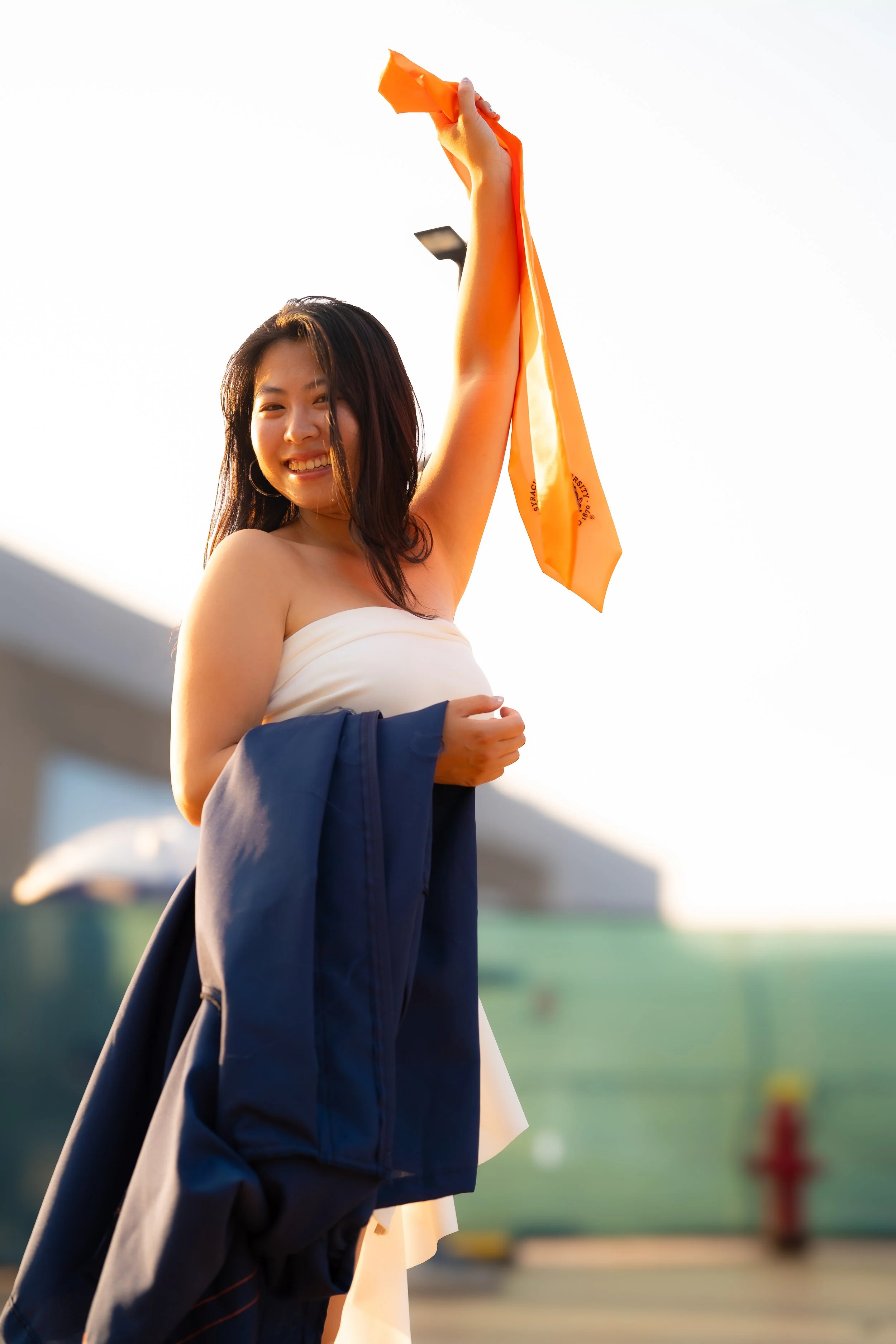 Young woman smiling, holding an orange object above her head, outdoors in a sports or school setting, with a blurry background including a green field, a building, and a red hydrant.