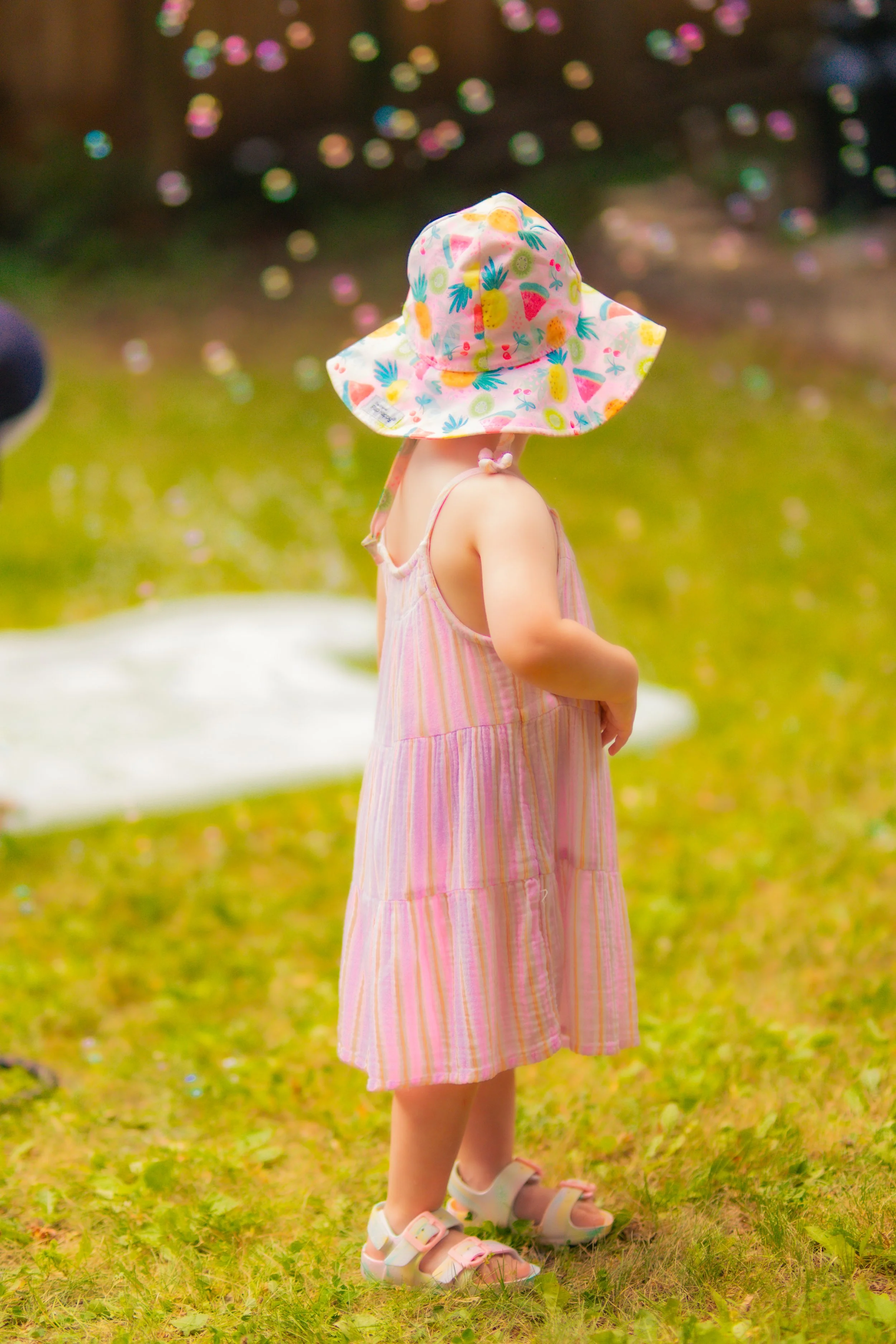 A young girl wearing a pink and yellow striped dress, white sandals, and a colorful wide-brimmed hat with fruit and tropical patterns, standing outdoors on grass with blurred colorful lights or reflections in the background.