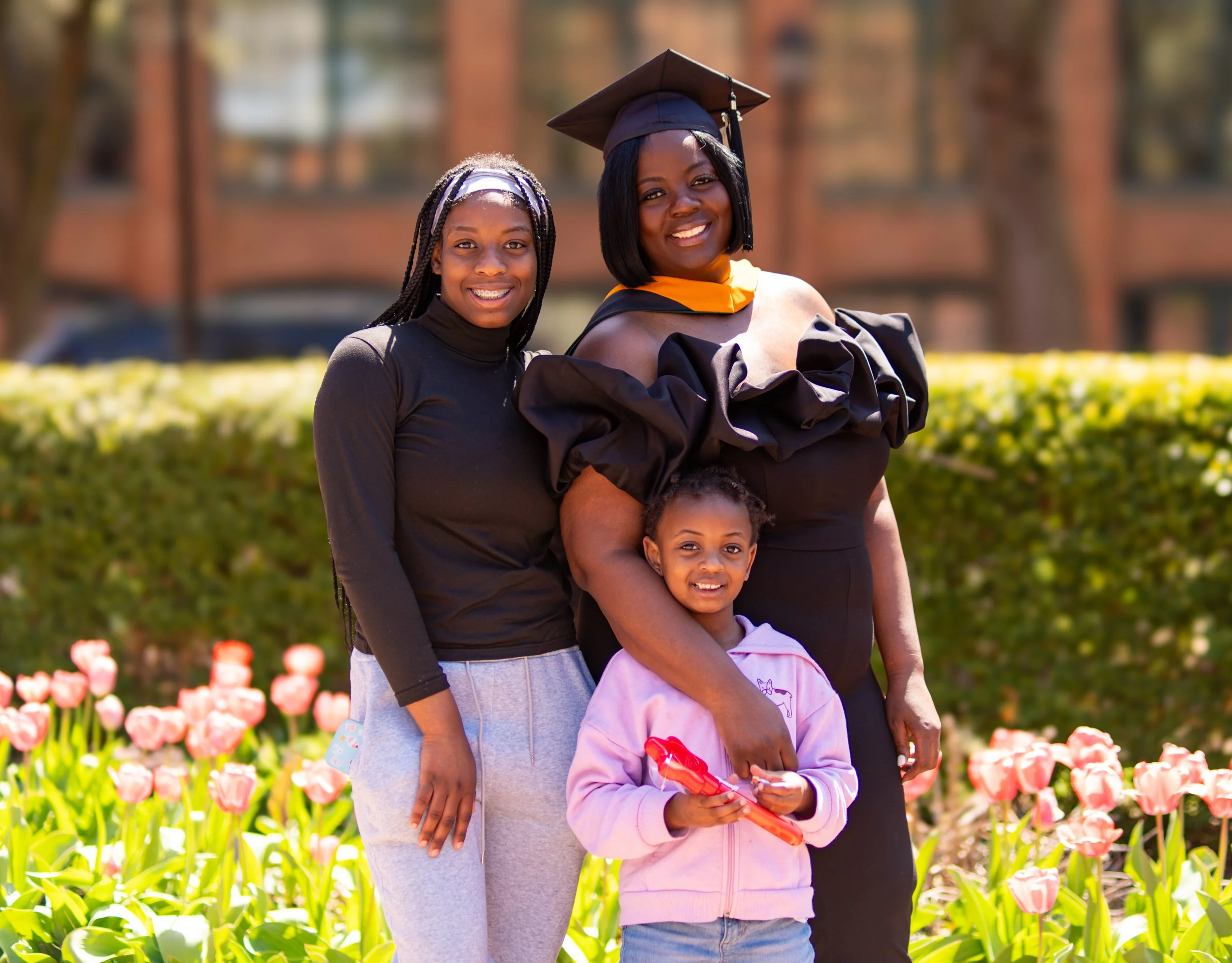 A group of three women and one girl standing outdoors in a garden with pink tulips, smiling for a photo.