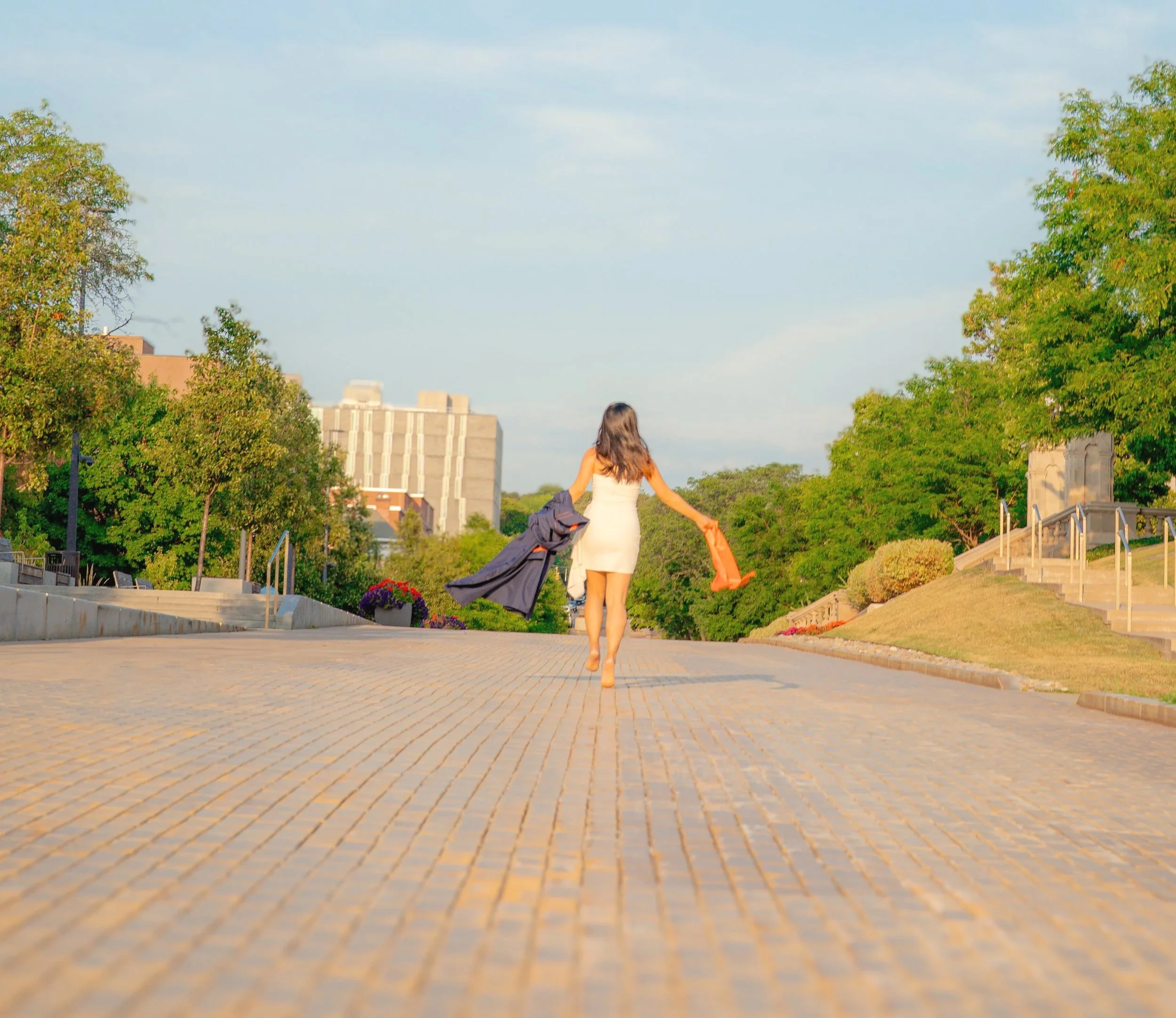 A woman in a white dress walking on a paved pathway in a park with green trees on either side and buildings in the background.