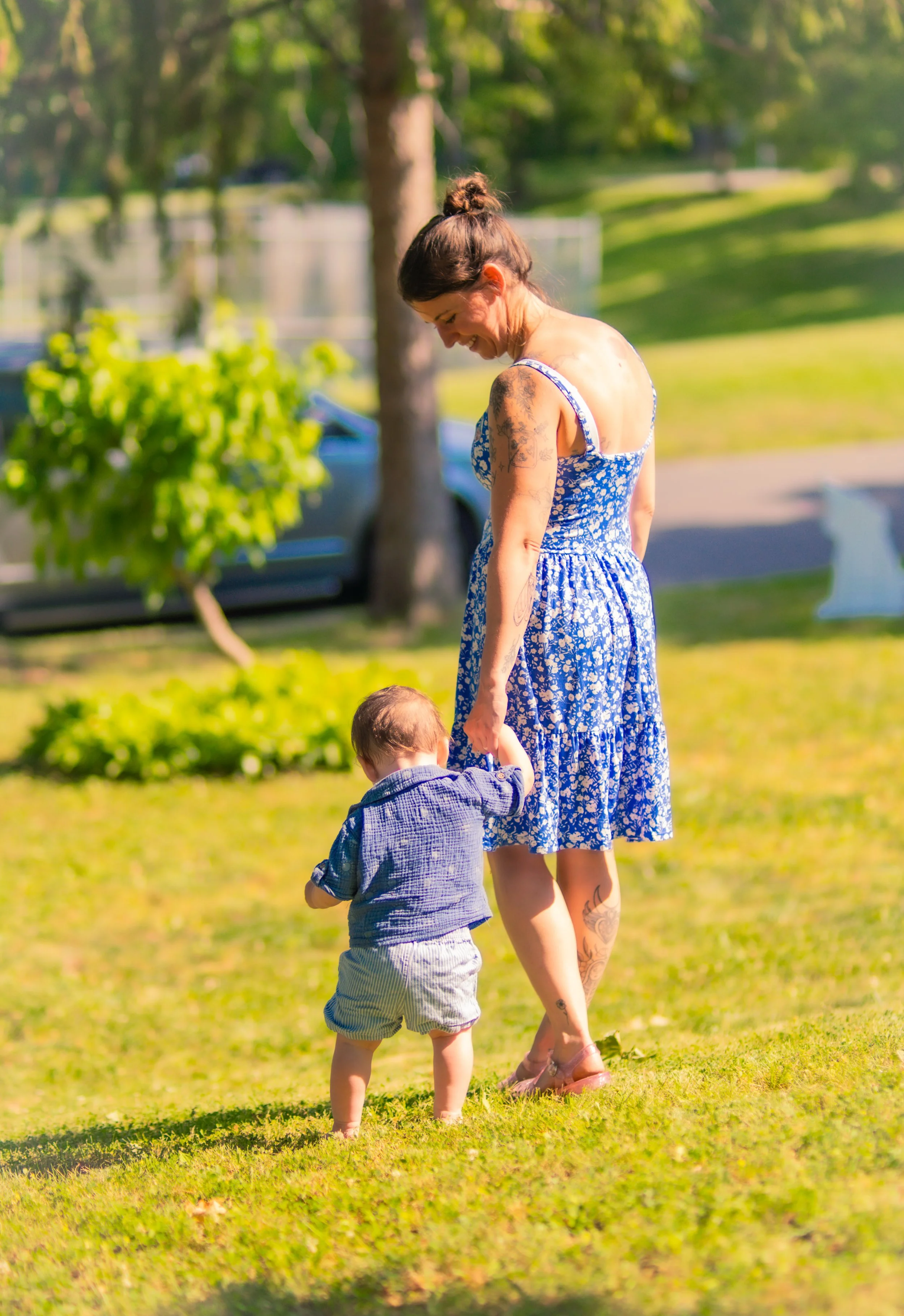 A woman with tattoos in a blue floral dress walking hand in hand with a young boy in a blue shirt outdoors in a park on a sunny day.