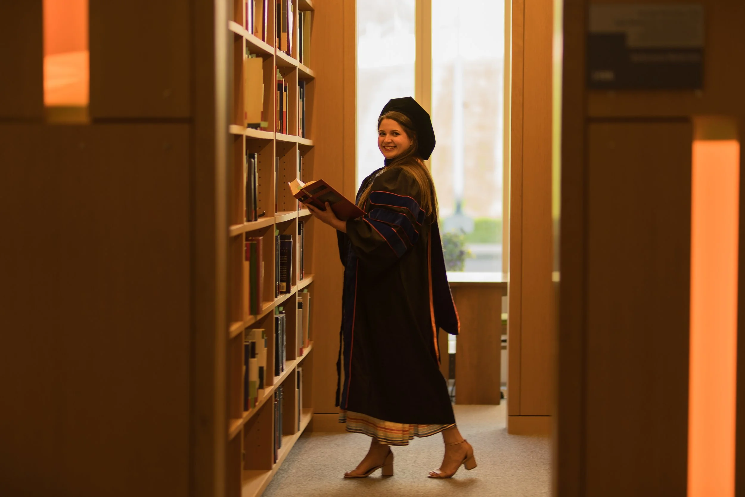 A young woman in a graduation gown and cap is browsing books on a library bookshelf, smiling and looking back at the camera.