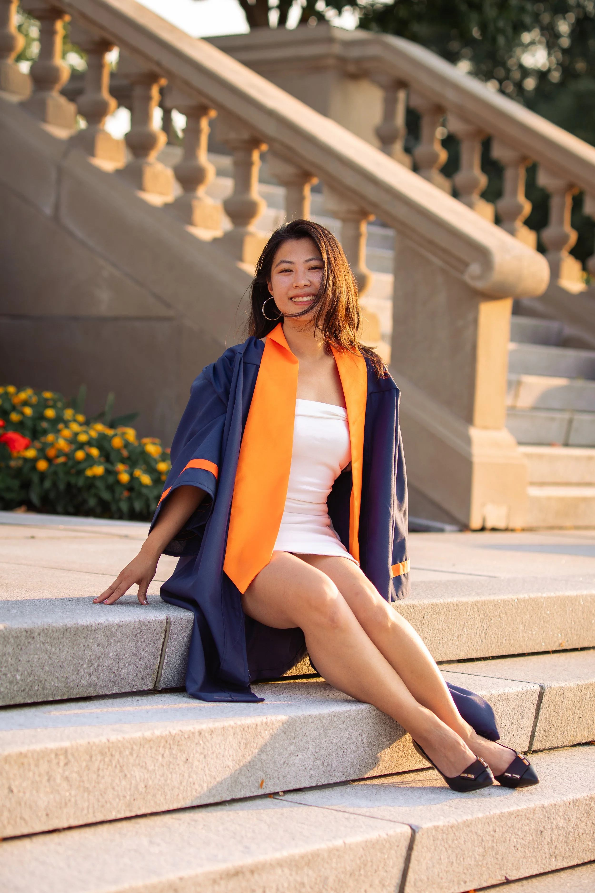 A young woman in a graduation gown sitting on steps outside, smiling at the camera, with flowers and a staircase in the background.