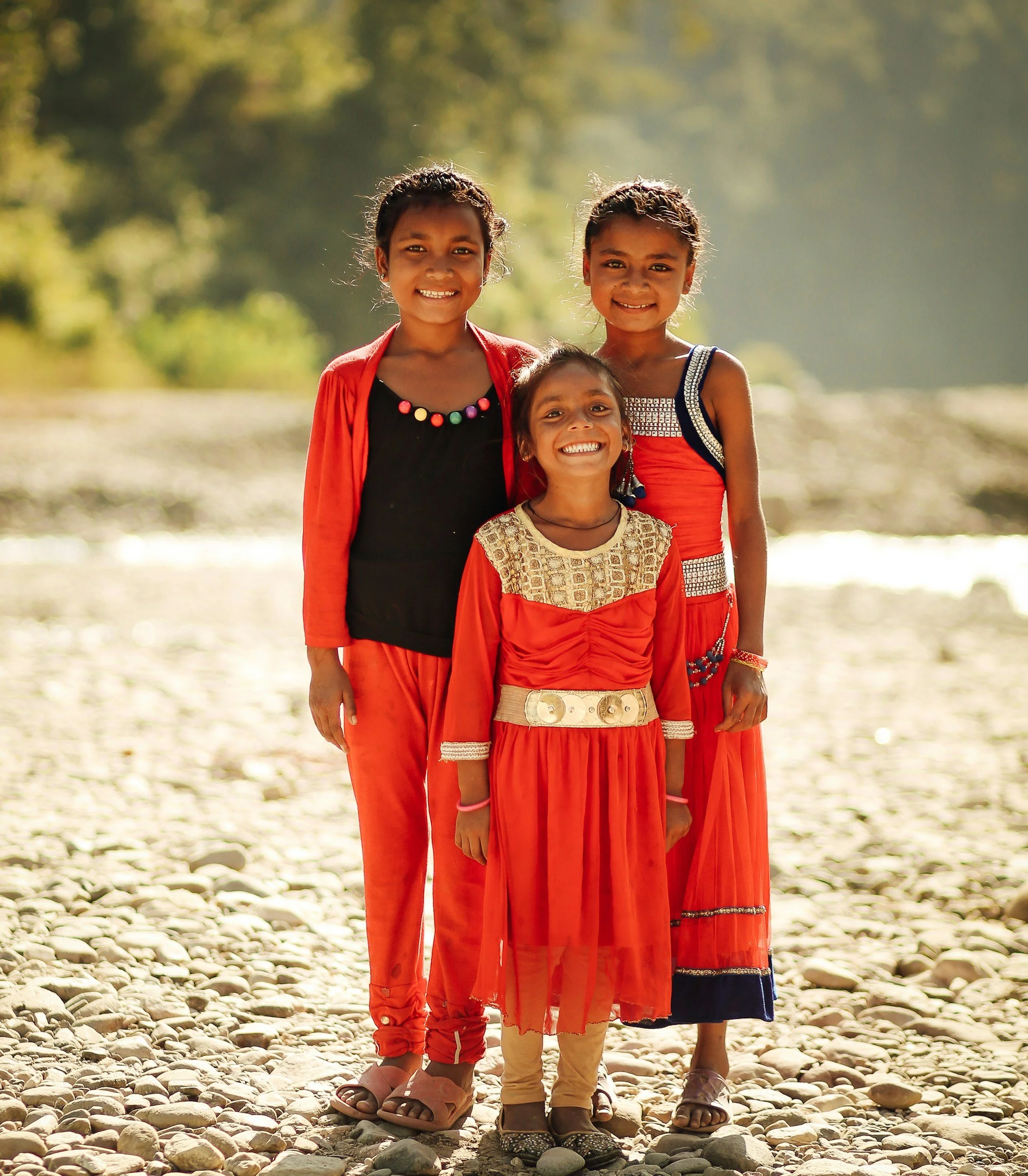 Four young girls smiling and standing together on a rocky surface outdoors, with a blurred green background and sunlight shining on them.