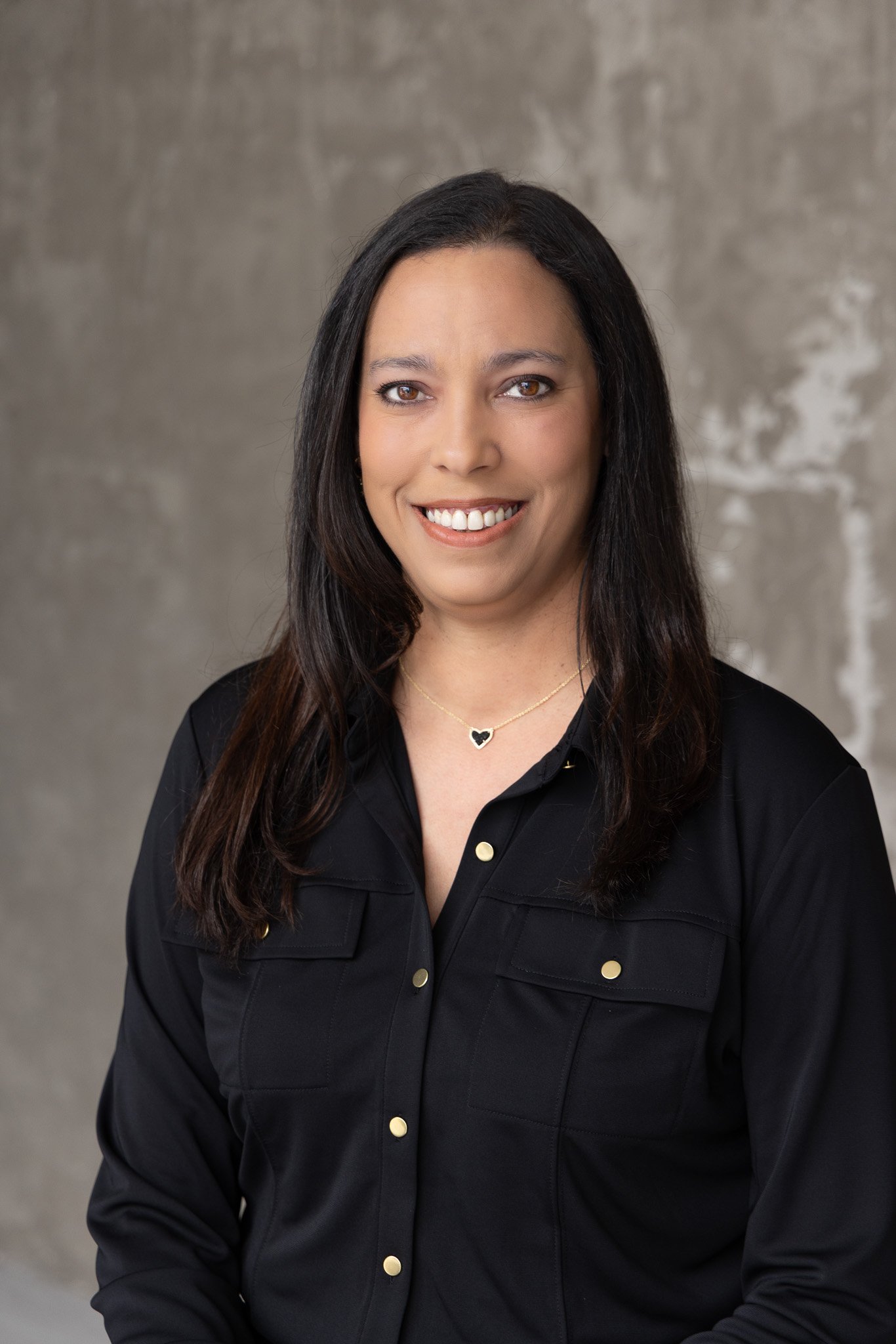 Portrait of a woman with long dark hair and light brown eyes, smiling, wearing a black shirt with gold buttons and a heart-shaped necklace, standing against a neutral gray textured background.