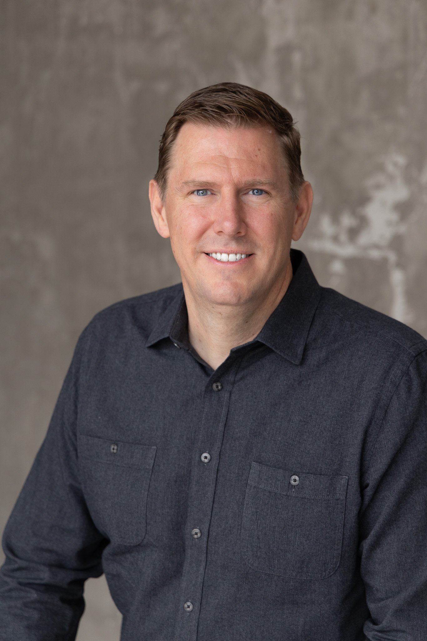 Portrait of a middle-aged man with short brown hair, blue eyes, and a friendly smile, wearing a dark gray button-up shirt, against a textured gray background.