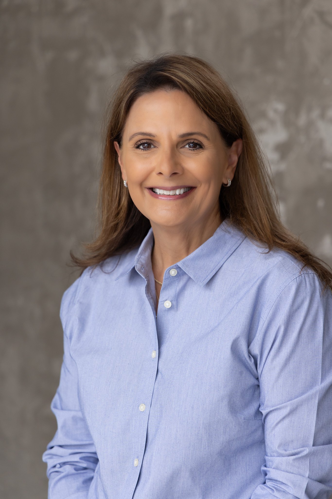 A woman with shoulder length brown hair, wearing a light blue button-up shirt, smiling, with a gray textured background.