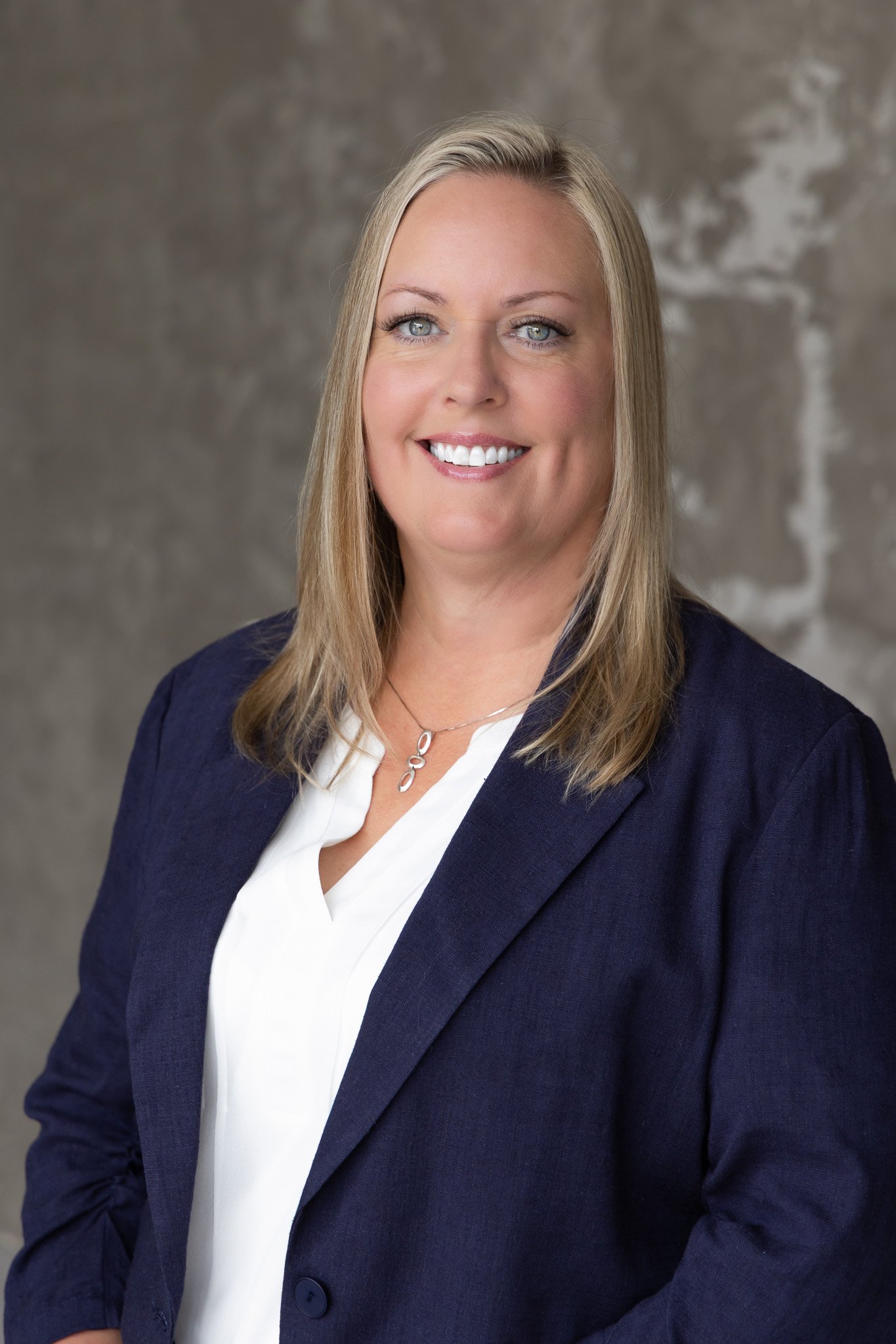 A smiling woman with blonde hair, wearing a navy blazer and a white blouse, standing against a textured gray background.