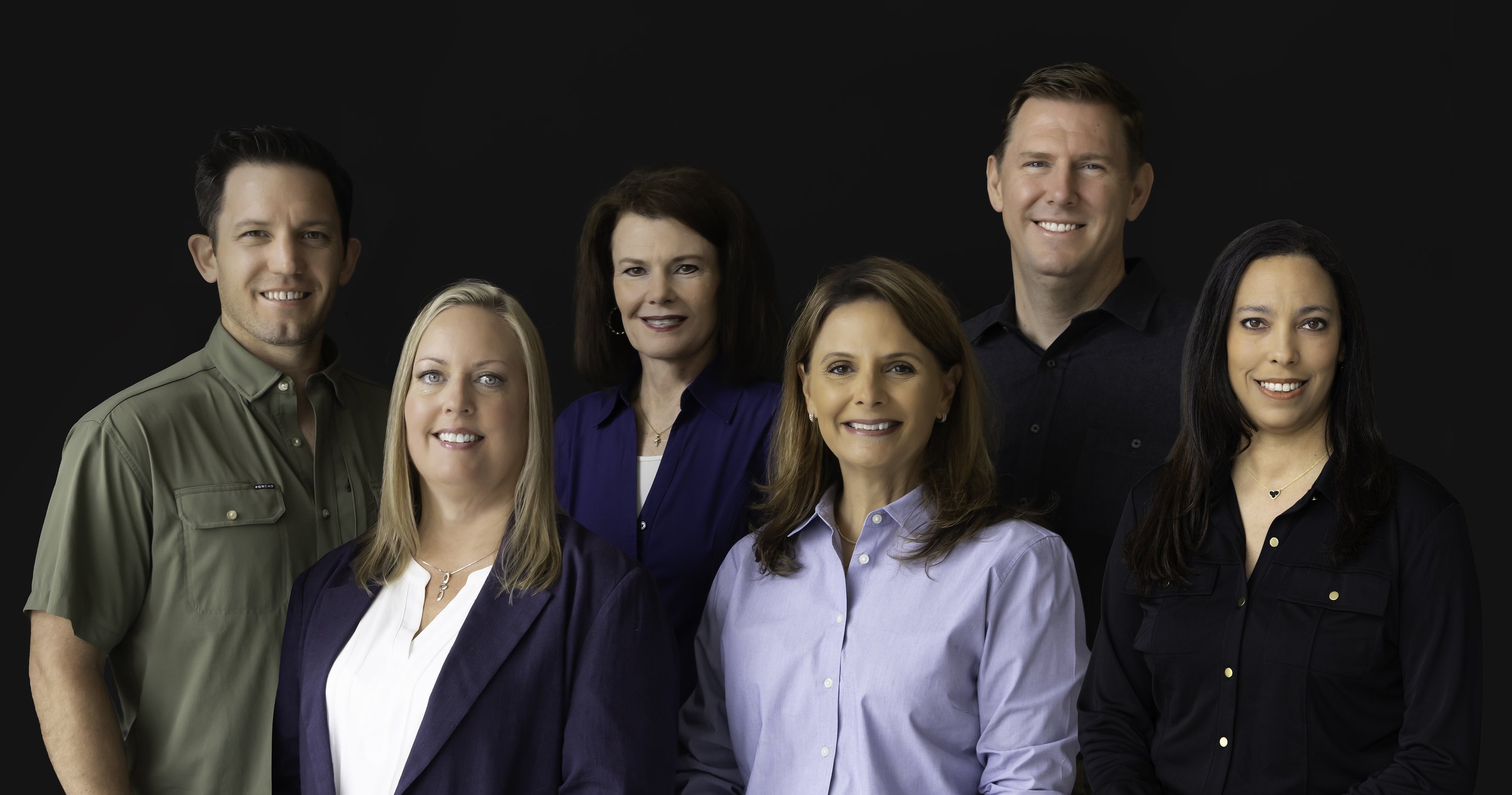 Group portrait of seven professionally dressed men and women smiling against a dark background.