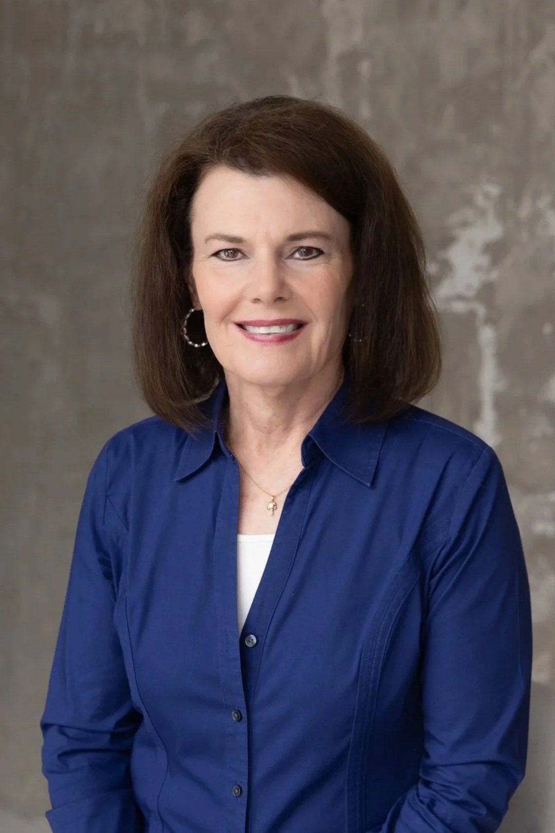 A smiling middle-aged woman with shoulder-length brown hair, wearing hoop earrings, a gold necklace with a cross, a blue button-up shirt, and a white top underneath, standing against a textured gray background.