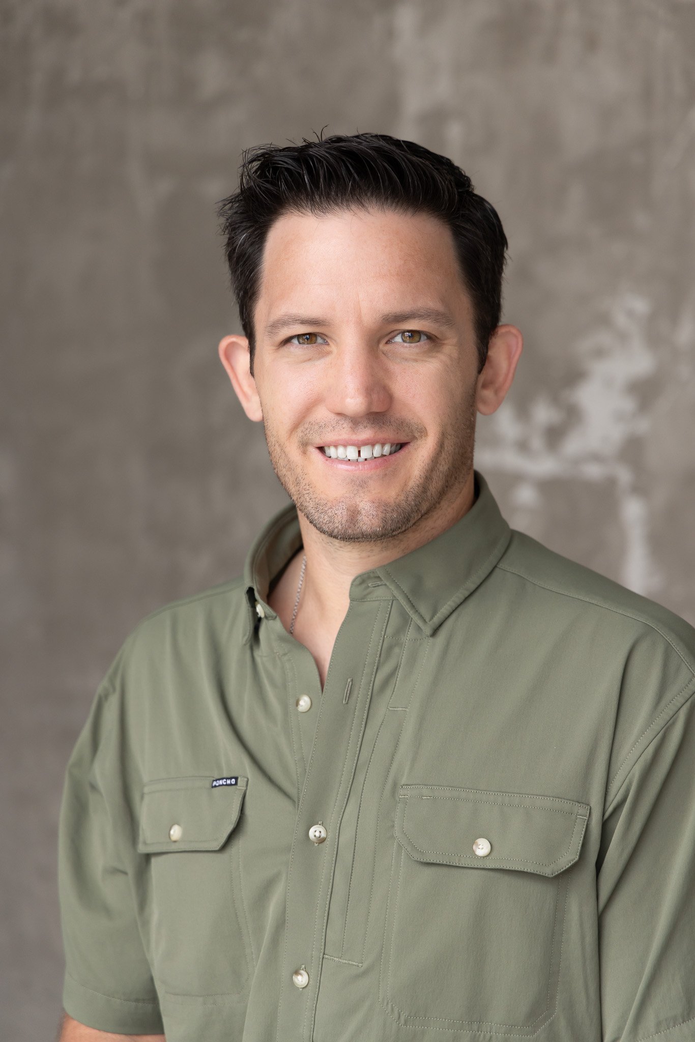 Head and shoulders photo of a smiling man with short dark hair, wearing an olive-green uniform with a name tag that says 'Pincho,' against a gray background.