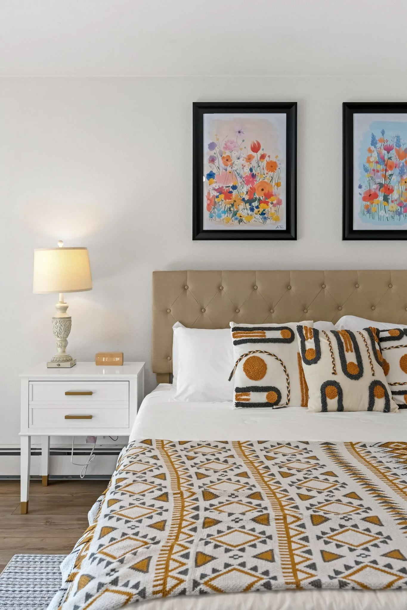 A bedroom with a bed, decorative pillows with geometric patterns, a beige tufted headboard, a white side table with a lamp, framed floral artwork on the wall, and a patterned bedspread.