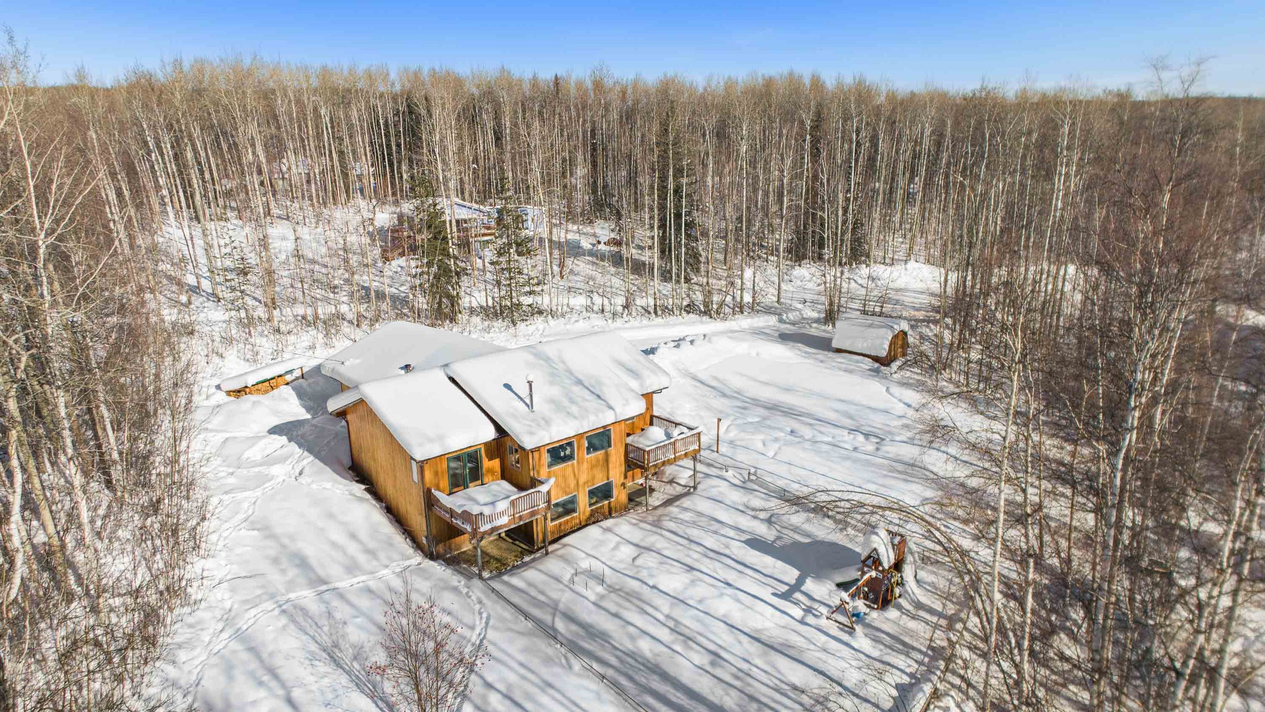 A wooden house with snow-covered roof and deck in a snowy landscape, surrounded by leafless trees and a dense forest in the background, under a clear blue sky.