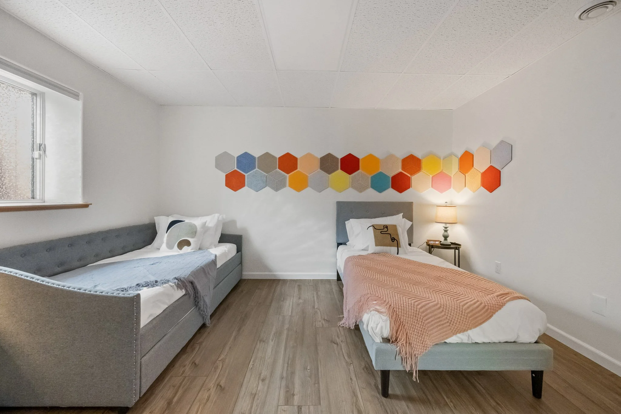 A bedroom with two beds, a gray upholstered bed on the left and a white bed on the right, with a colorful hexagonal wall art piece, a window on the left, a nightstand with a lamp on the right, and wooden flooring.
