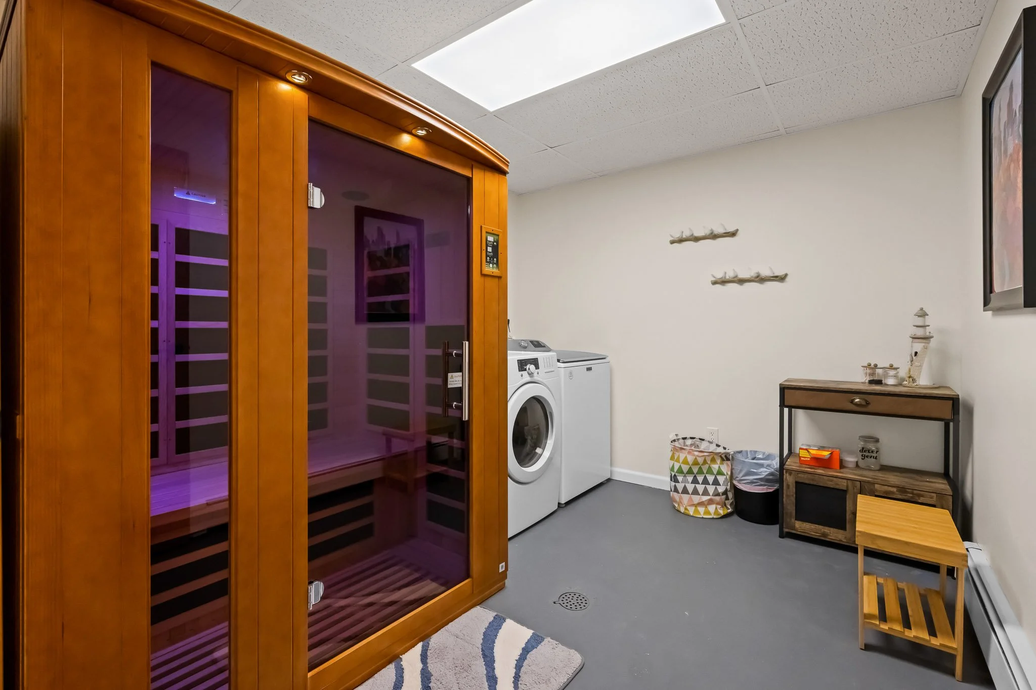 A room with a wooden sauna, washing machine, laundry basket, small wooden table, and wall decorations.