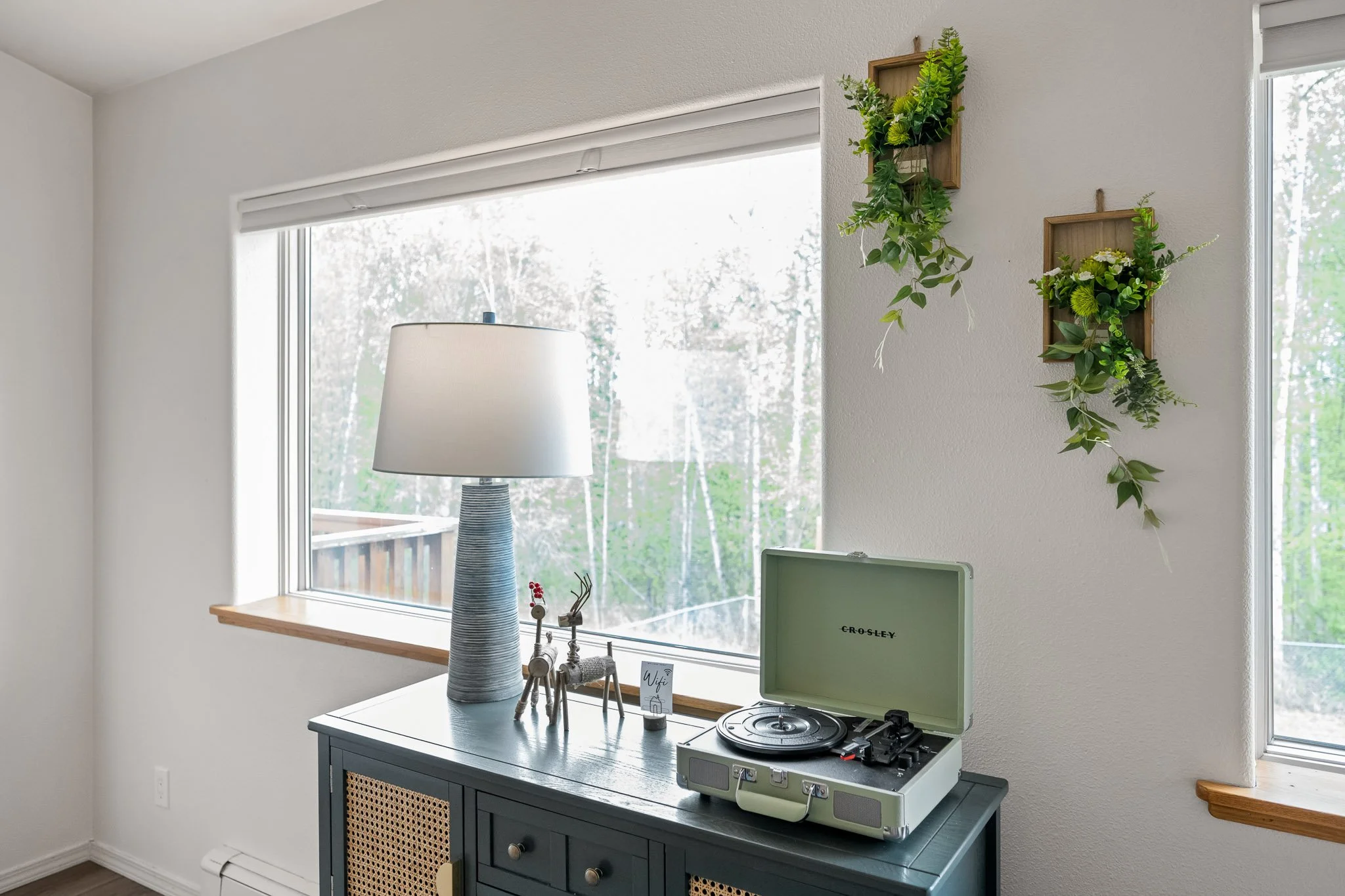 A corner of a room with a large window, a lamp with a flat base and a cylindrical shade, a blue cabinet with a small drawer, and a vintage portable record player on top. Two small reindeer and a Wi-Fi sign are also on the cabinet. On the wall above i
