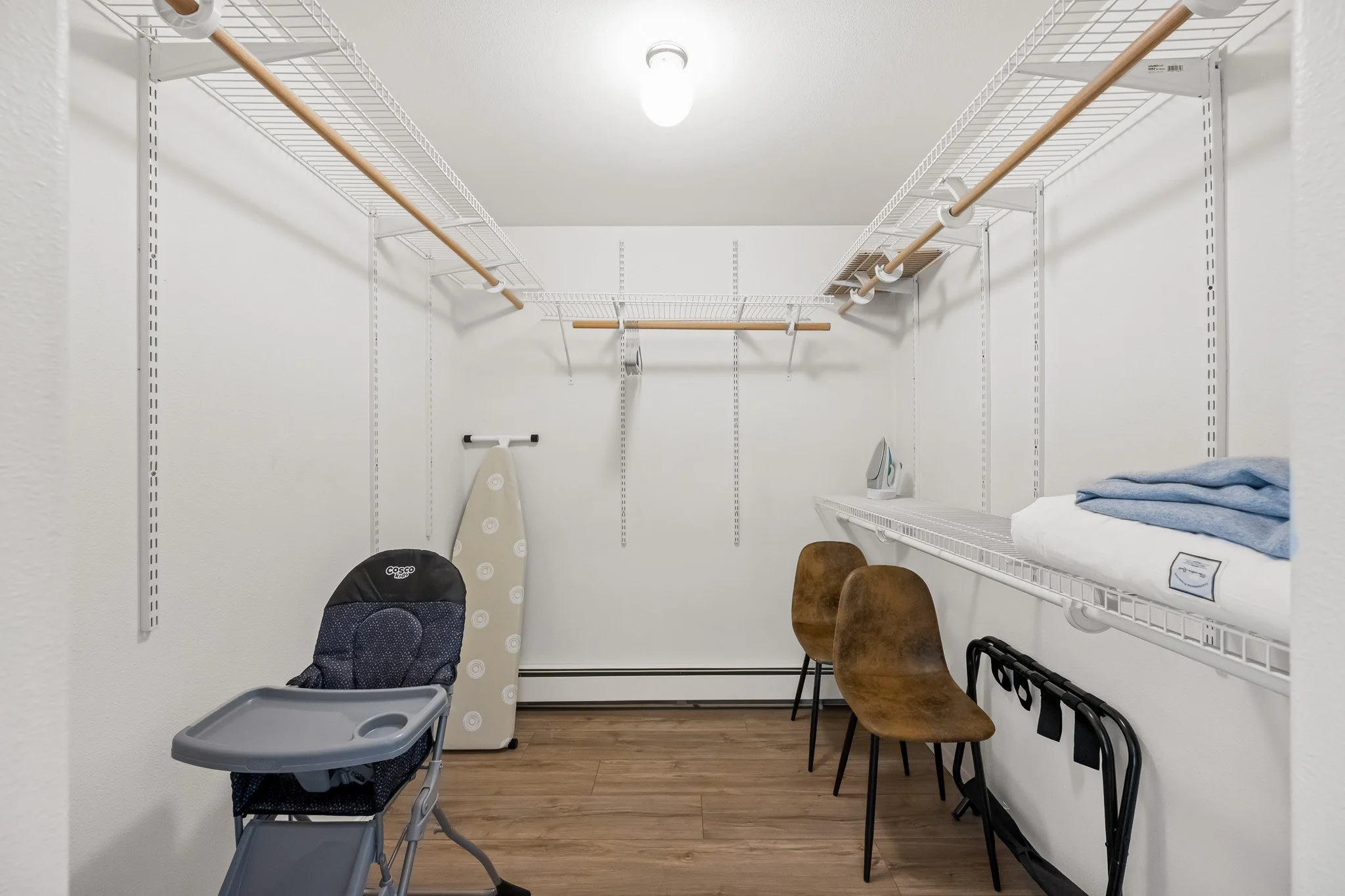 A walk-in closet with white wire shelving and wooden rod bars for hanging clothes. There is an iron resting on the shelf, an ironing board, two wooden chairs, a black and blue baby stroller, and a high chair, all placed against white walls and a wood
