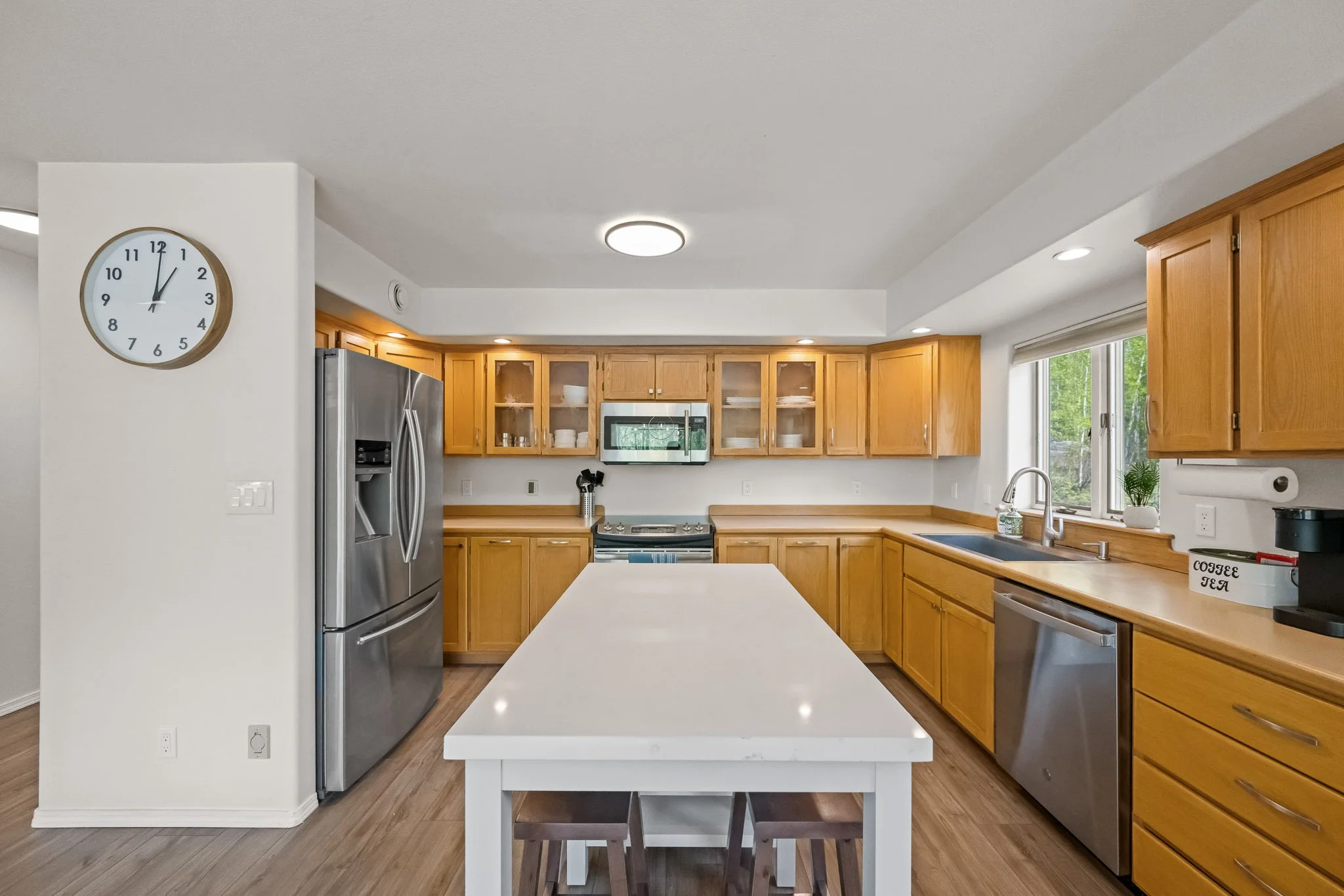Kitchen with wooden cabinets, stainless steel appliances, a white island in the center, and a large window with a view of greenery.