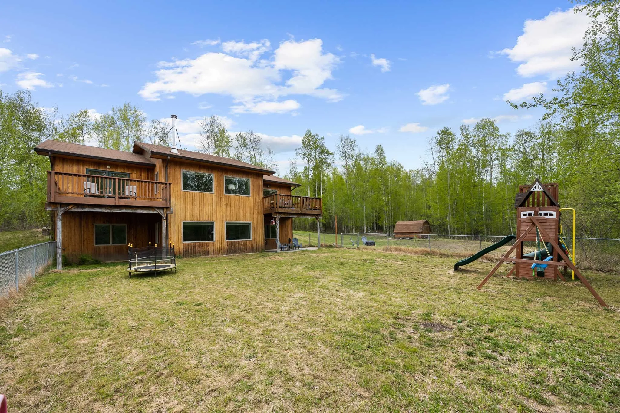 A backyard scene with a wooden house featuring a balcony, a fenced yard with a small trampoline, and a playset with a slide, surrounded by green trees and a clear blue sky with scattered clouds.