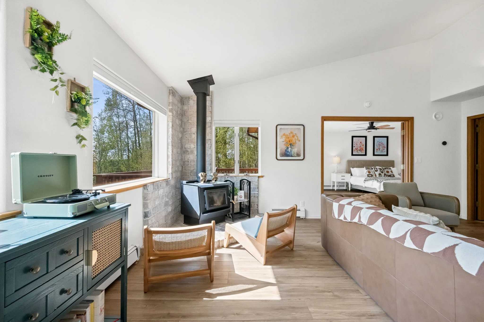 A bright living room with large windows, a wood stove, framed artwork, a beige couch with a patterned blanket, a blue cabinet with a turntable on top, and green plants on the wall.