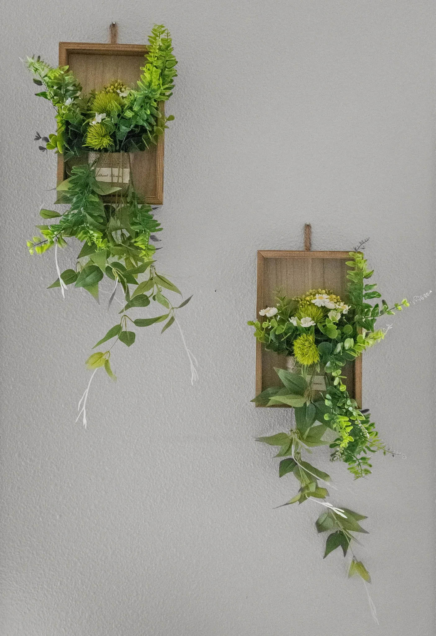 Two wall-mounted wooden frames with artificial green foliage and small white and yellow flowers, hanging on a beige wall.