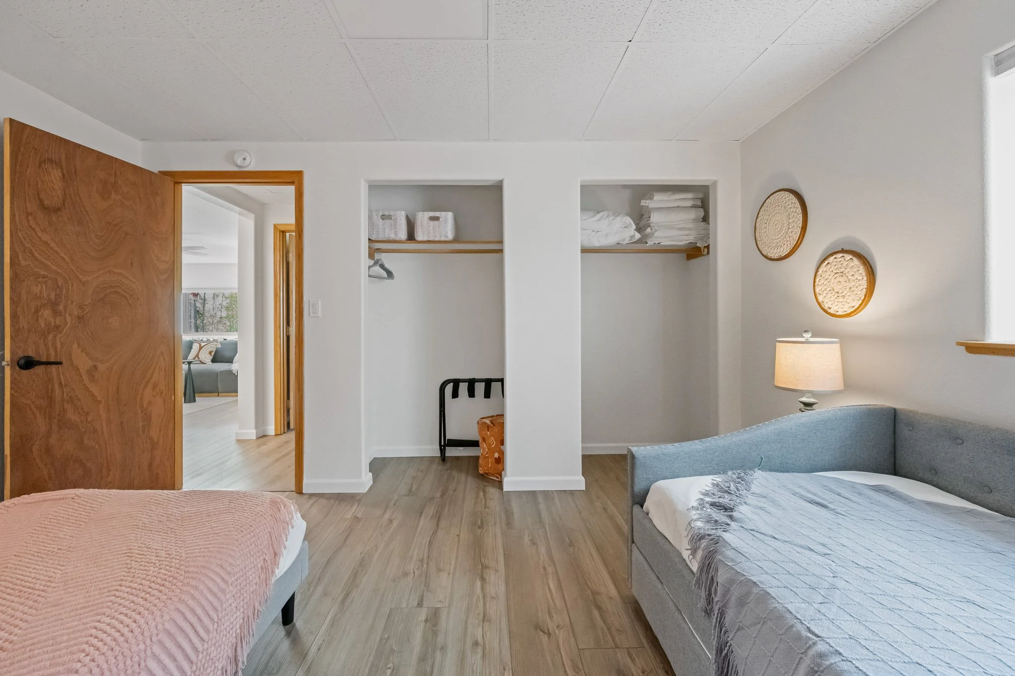 View of a modern bedroom with a white wall, two beds with pink and gray bedding, a wooden door, built-in shelves with folded towels, wall art, a table lamp, and a glimpse into a living room with a sofa.