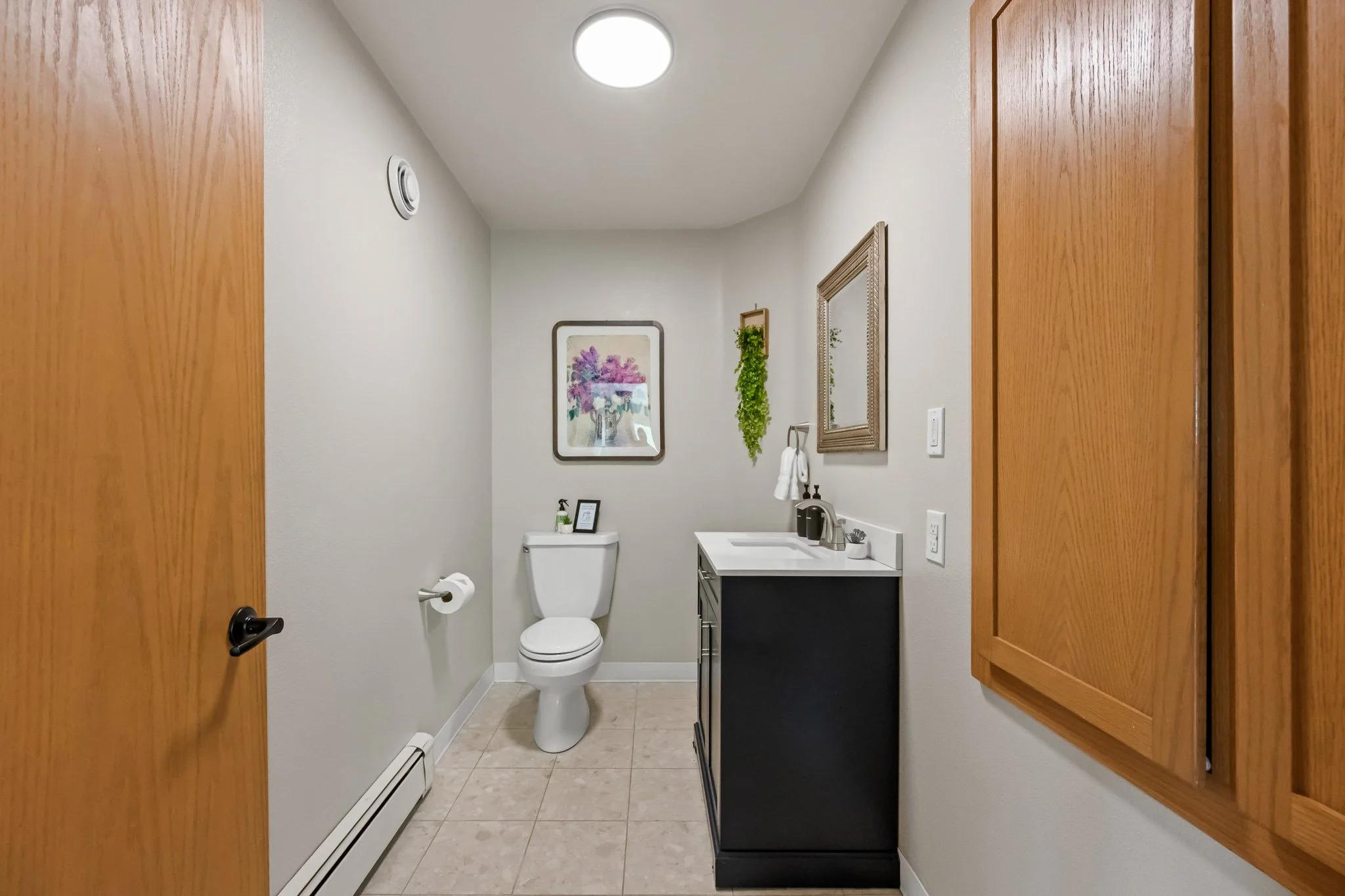 A small bathroom with a white toilet, a black vanity with a white sink, wall art, a mirror, and wall decor. Wooden cabinets flank the doorway.