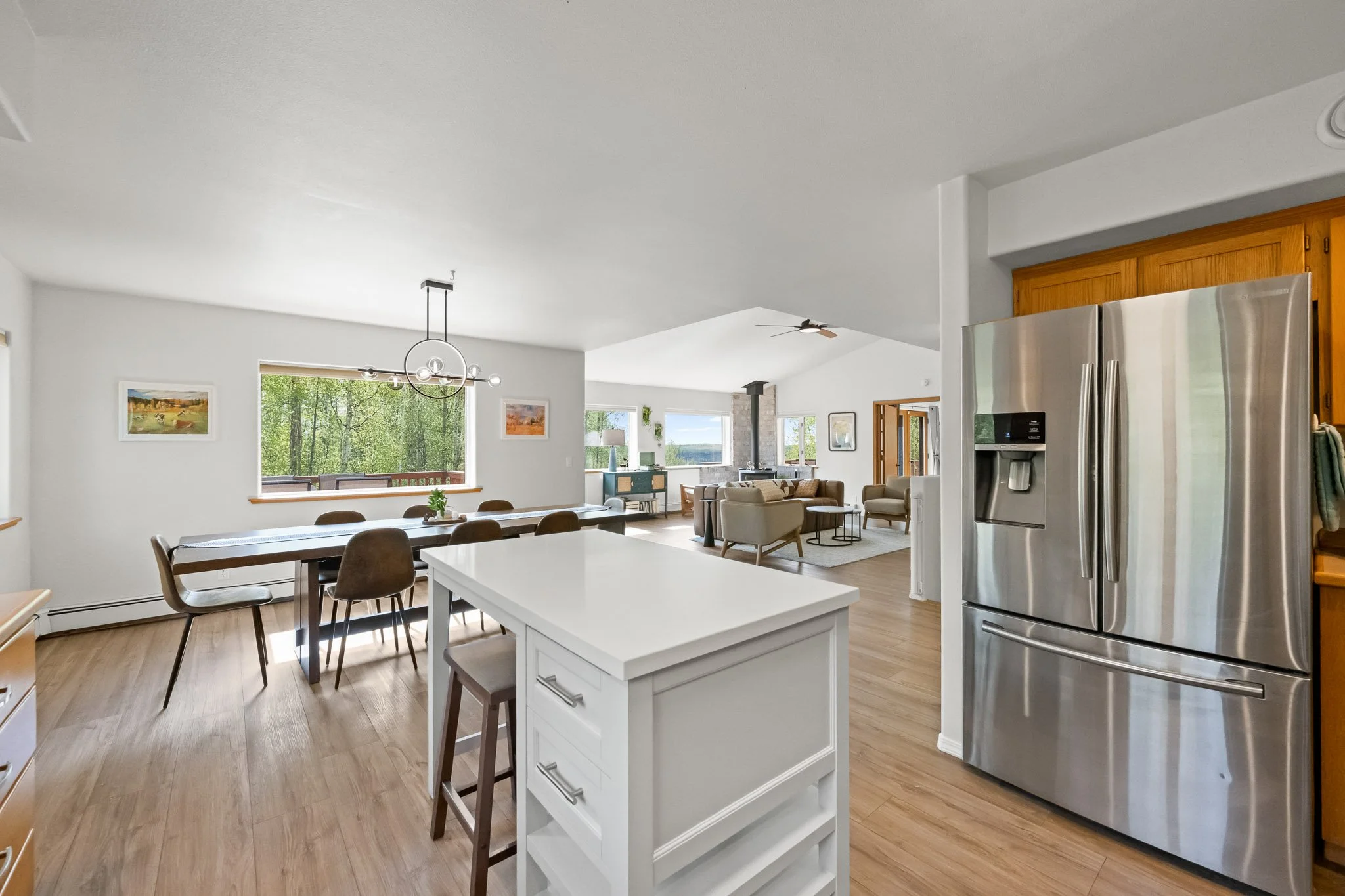 Open-concept kitchen and living room with large windows, modern chandelier, and stainless steel refrigerator.