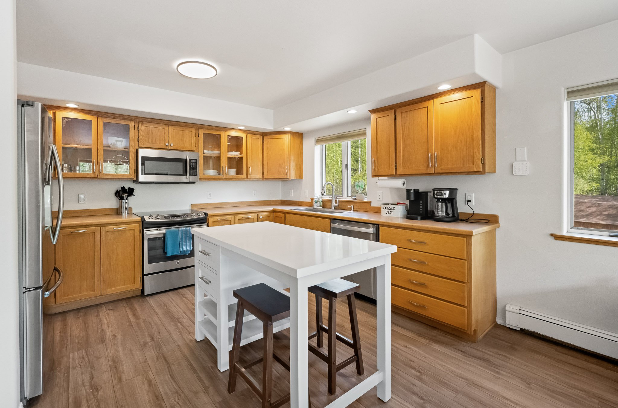 A kitchen with light wood cabinets, stainless steel appliances, a white island, and two wooden bar stools near a window showing trees outside.
