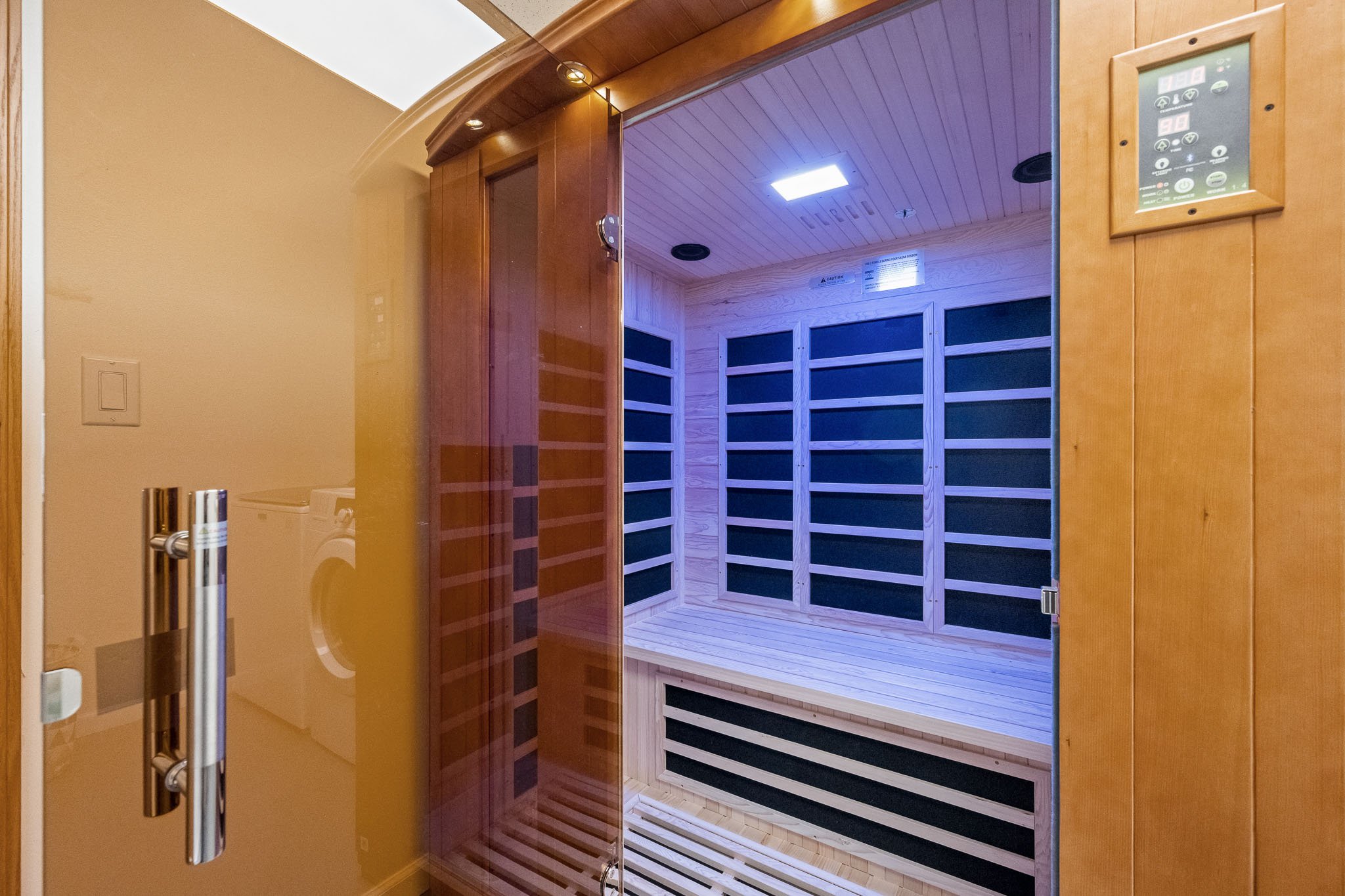 Image of a modern cedar sauna room with wooden bench seating and a faux wood panel ceiling, seen through a partially open glass door with a metal handle.