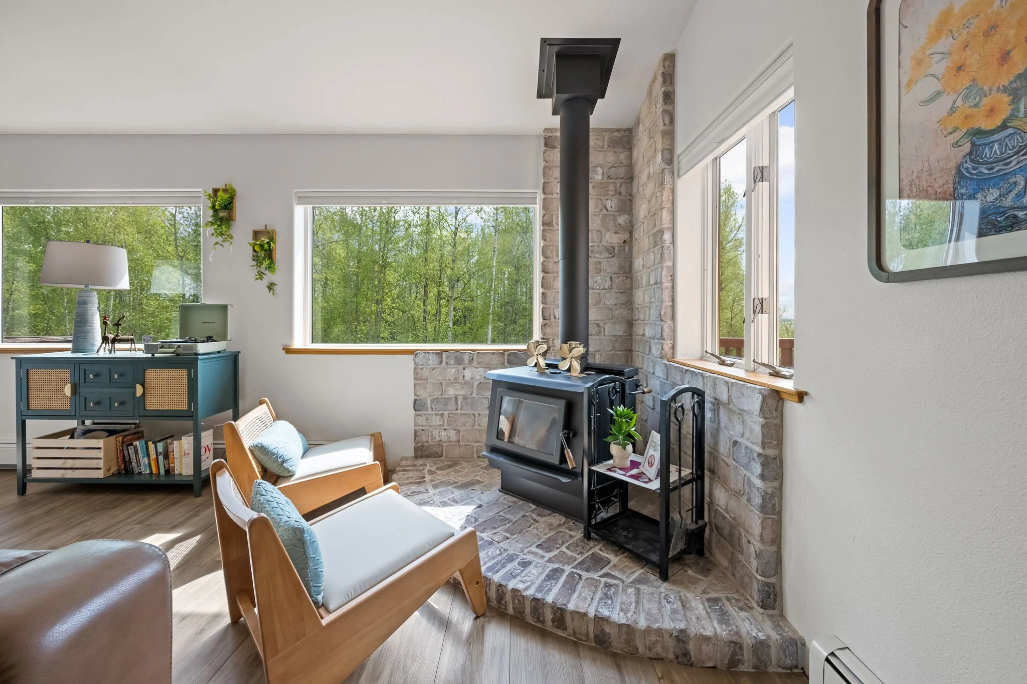 Living room with large windows showing green trees, a brick fireplace with a wood stove, a blue sideboard, and modern armchairs with blue cushions.