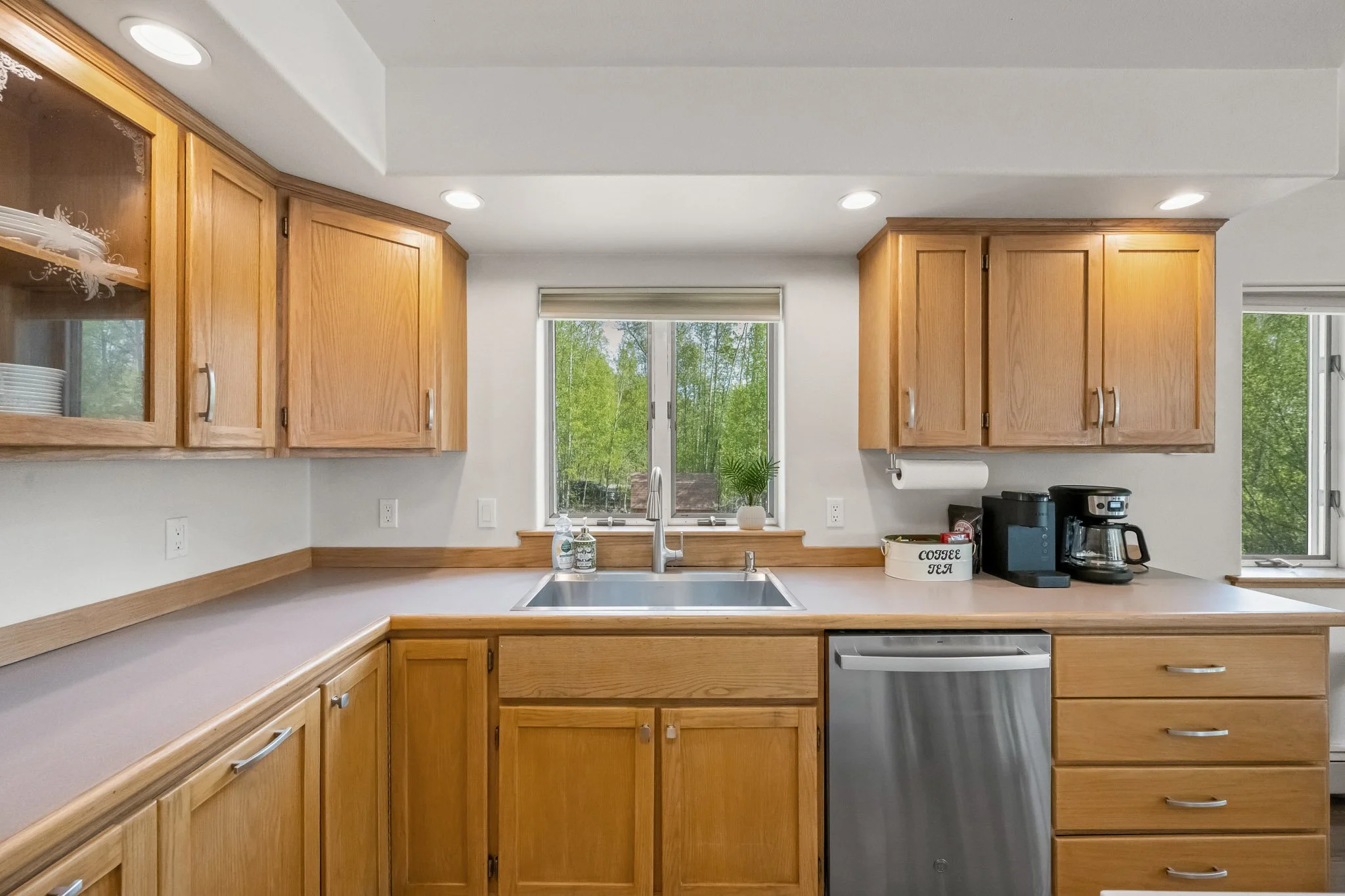 Kitchen with wooden cabinets, white walls, large window, and countertop with coffee maker and tea container.