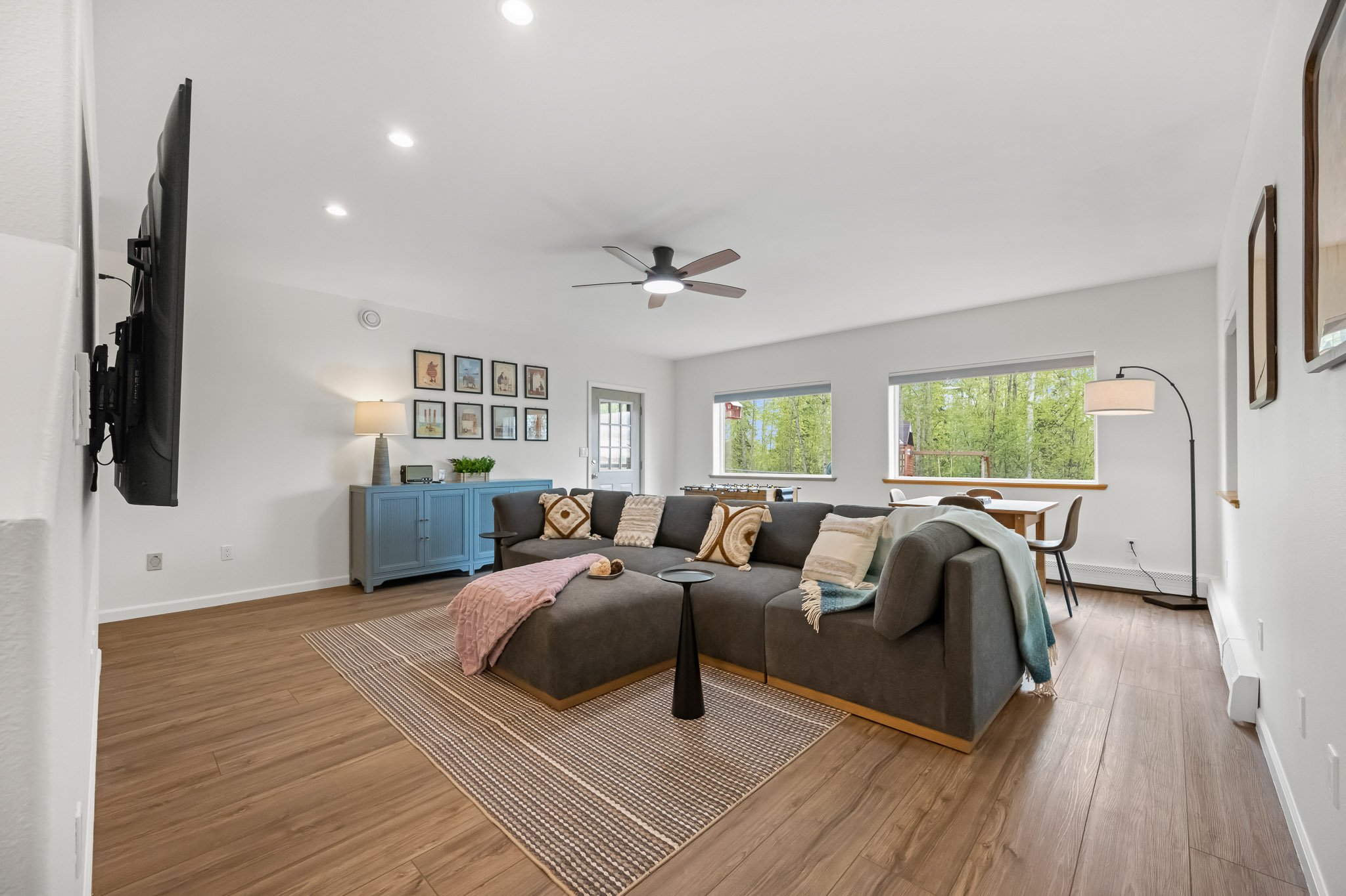 Living room with white walls, wooden flooring, a dark gray sectional sofa with pillows, a pink blanket, a blue sideboard with a lamp and plant, wall art, large windows showing green trees, a ceiling fan, and a lamp