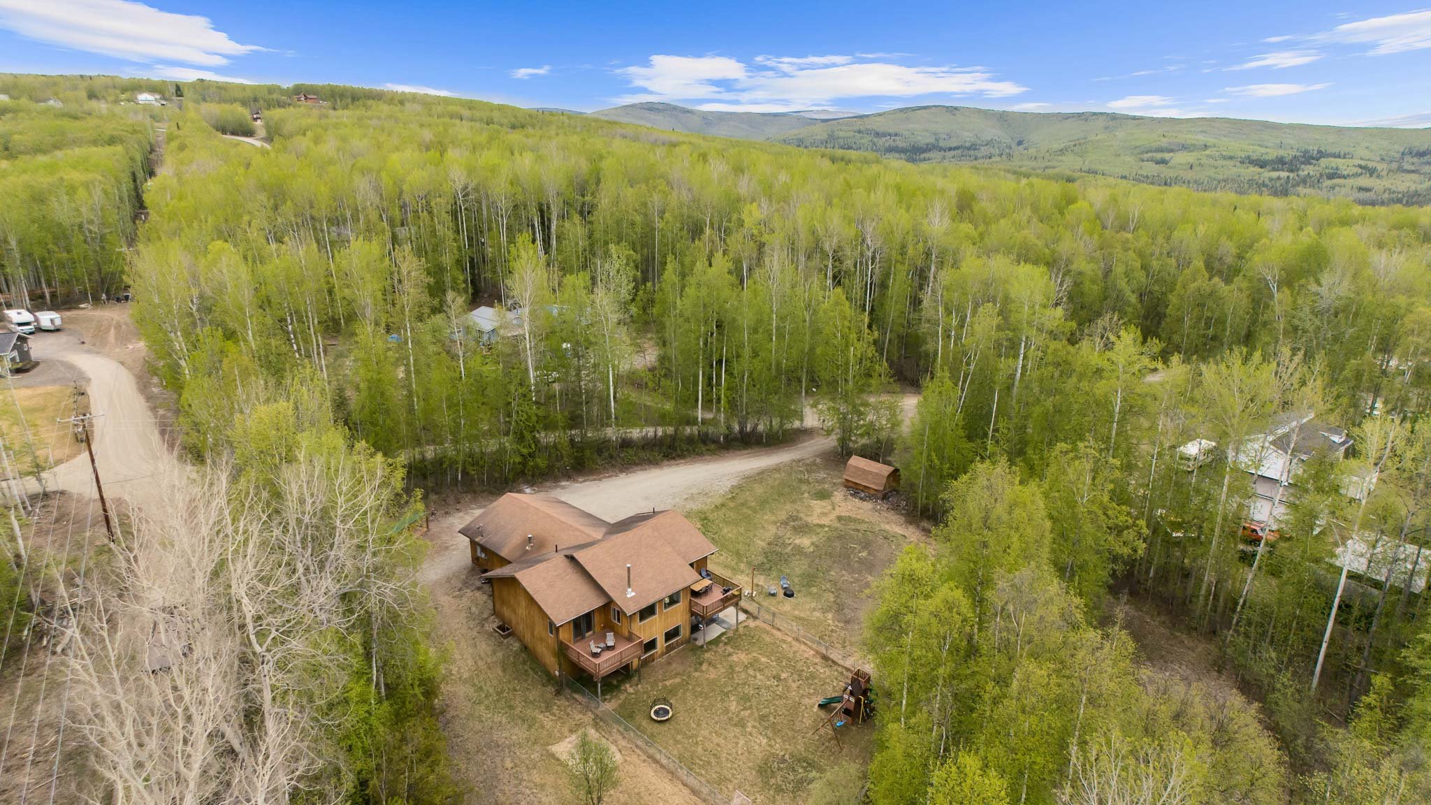 Aerial view of a house in a forested area with surrounding trees, dirt roads, and additional houses in the distance, set against a backdrop of distant hills and a blue sky with scattered clouds.