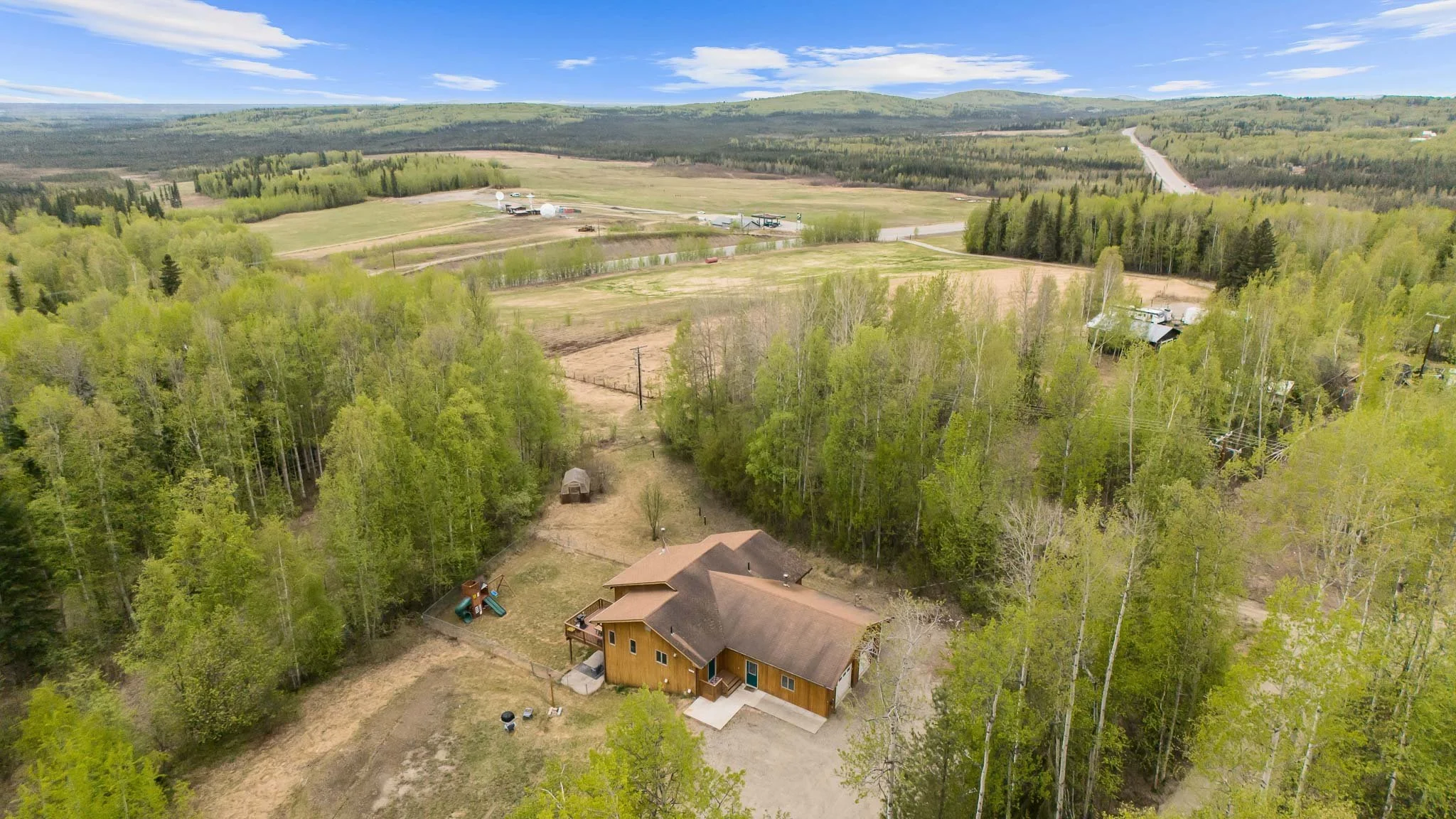 Aerial view of a rural landscape with houses, trees, fields, and distant hills under a partly cloudy sky.