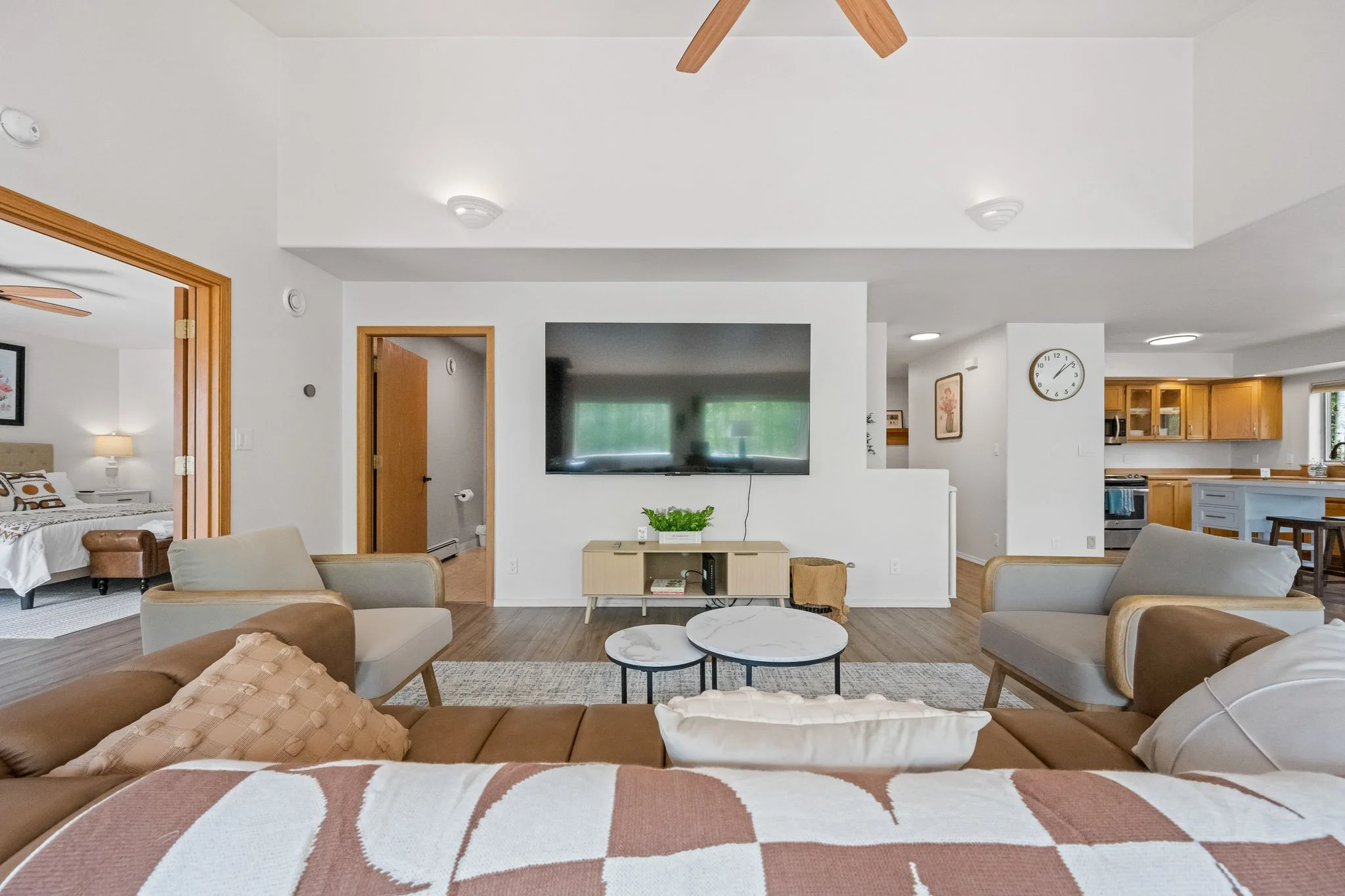 Interior of a modern living room with beige and white furniture, a wall-mounted TV, and wooden accents. Open doorway reveals a bedroom with a bed and nightstand. Kitchen with wooden cabinets and stainless steel appliances visible in the background.