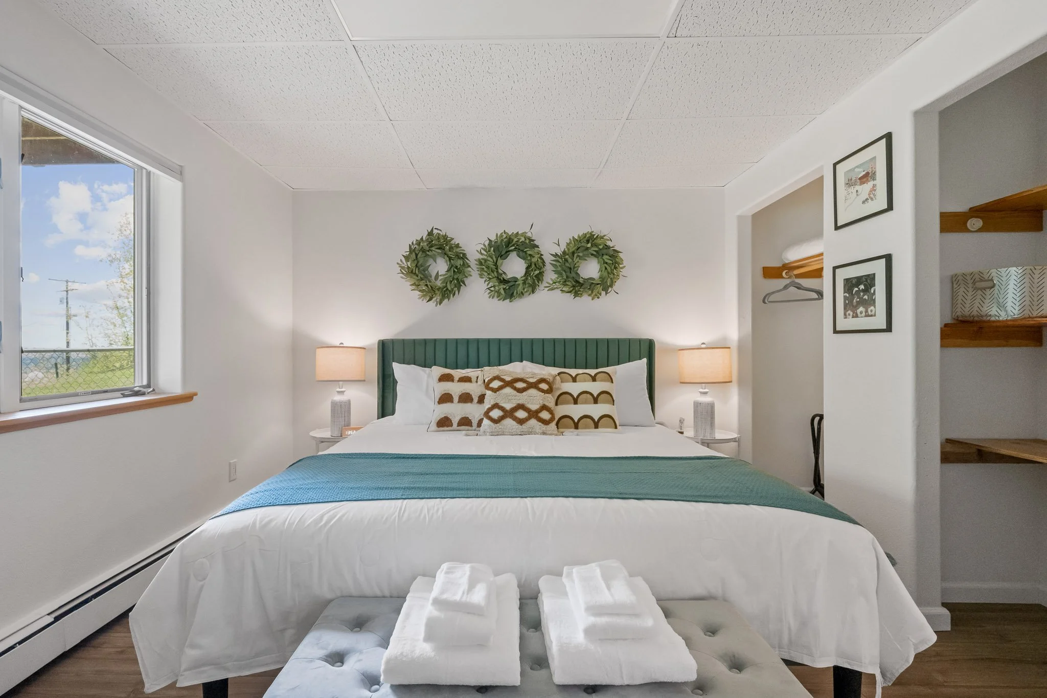 A cozy bedroom with a large bed, decorative pillows, green headboard, and two nightstands with lamps. Wreaths hang on the wall above the bed. There is a window on the left side with a view of blue sky, clouds, and trees outside. To the right, an open