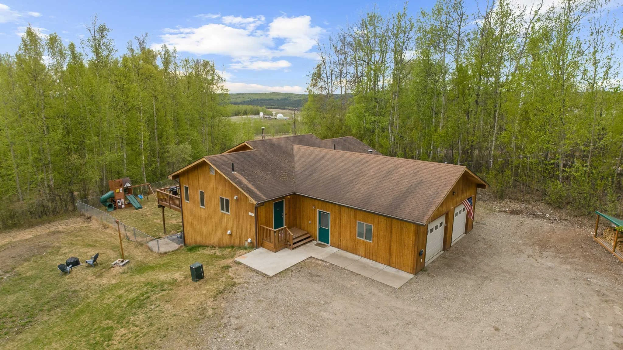 Aerial view of a wooden house with a sloped roof, part of a fenced backyard with playground equipment, and a green landscape with trees and hills in the background.