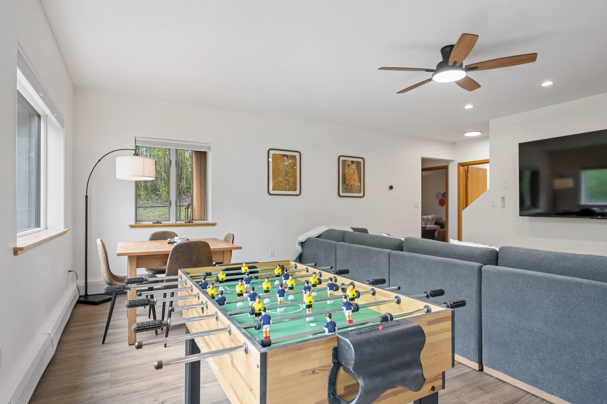 Living room with a foosball table, a gray sectional sofa, a dining table with four chairs, a standing lamp, a ceiling fan with wooden blades, a window showing outdoor greenery, and wall art.