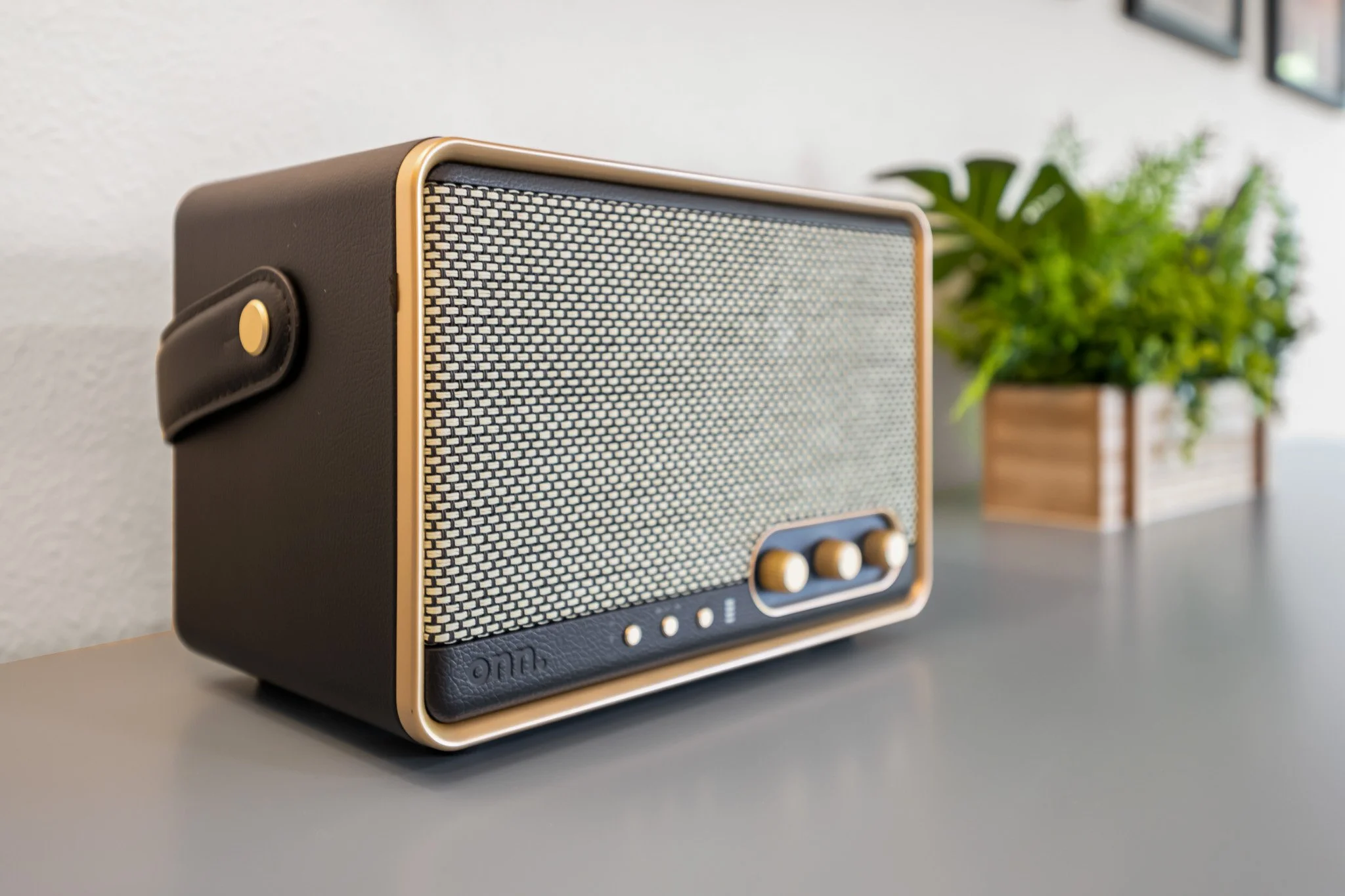 A vintage-style portable radio with a black body, cream-colored trim, and gold speaker grill, placed on a flat surface with a potted plant and framed pictures in the background.