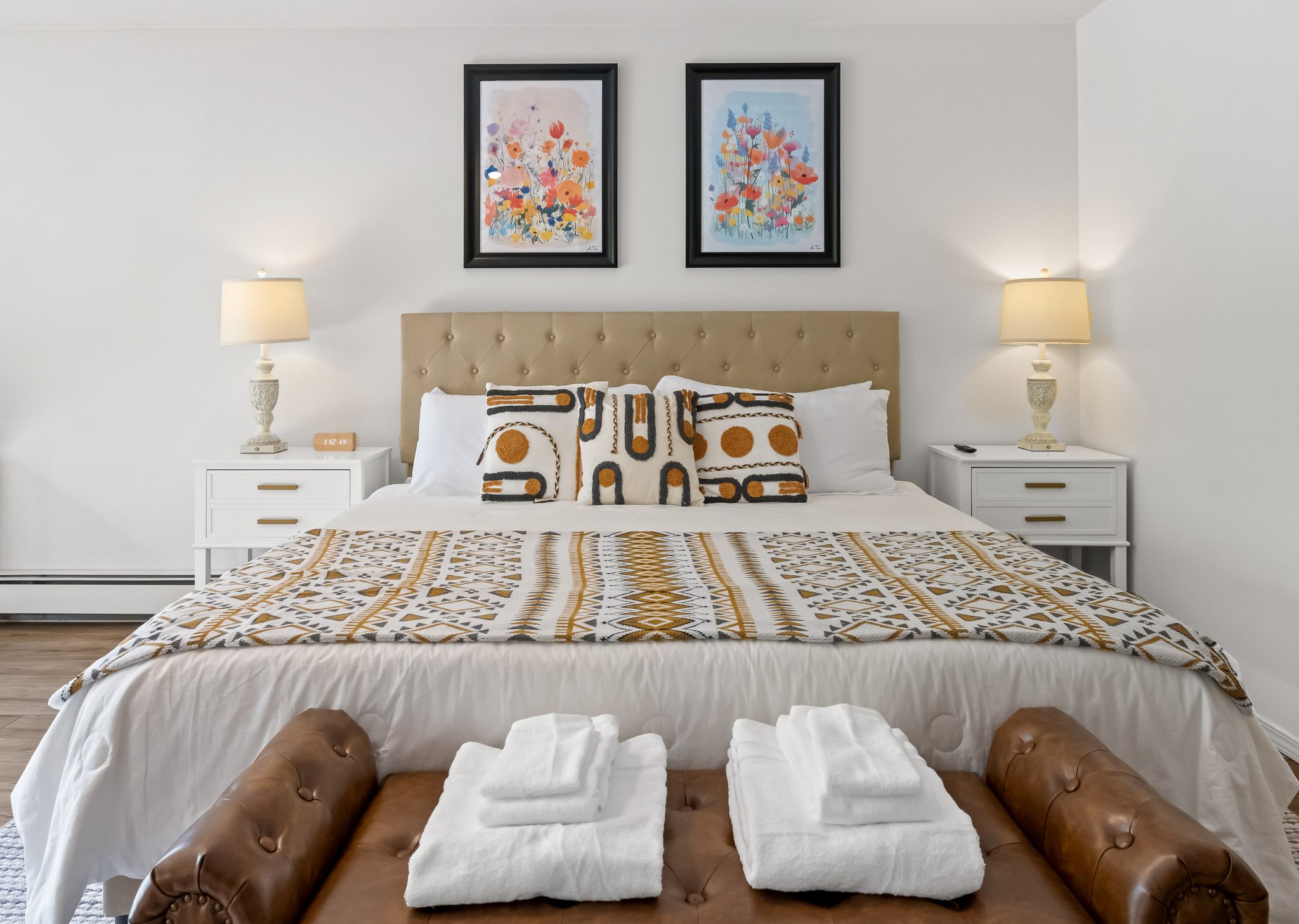A neatly made bed with white sheets, patterned throw pillows, and a quilted coverlet in a bedroom. Two white nightstands with lamps flank the bed, and two framed floral prints hang on the plain white wall above. A leather bench with rolled arms sits 