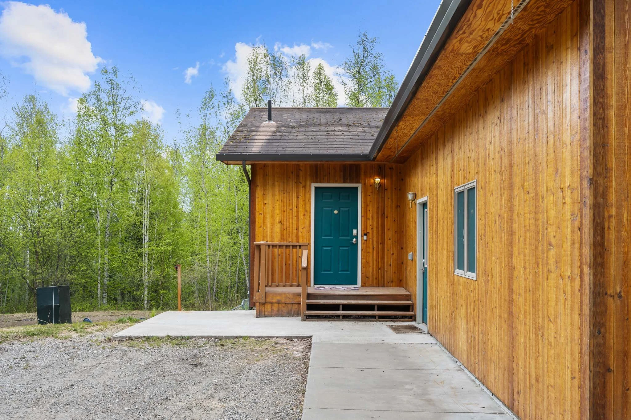 New wood house exterior with teal front door, surrounded by green trees and a blue sky.