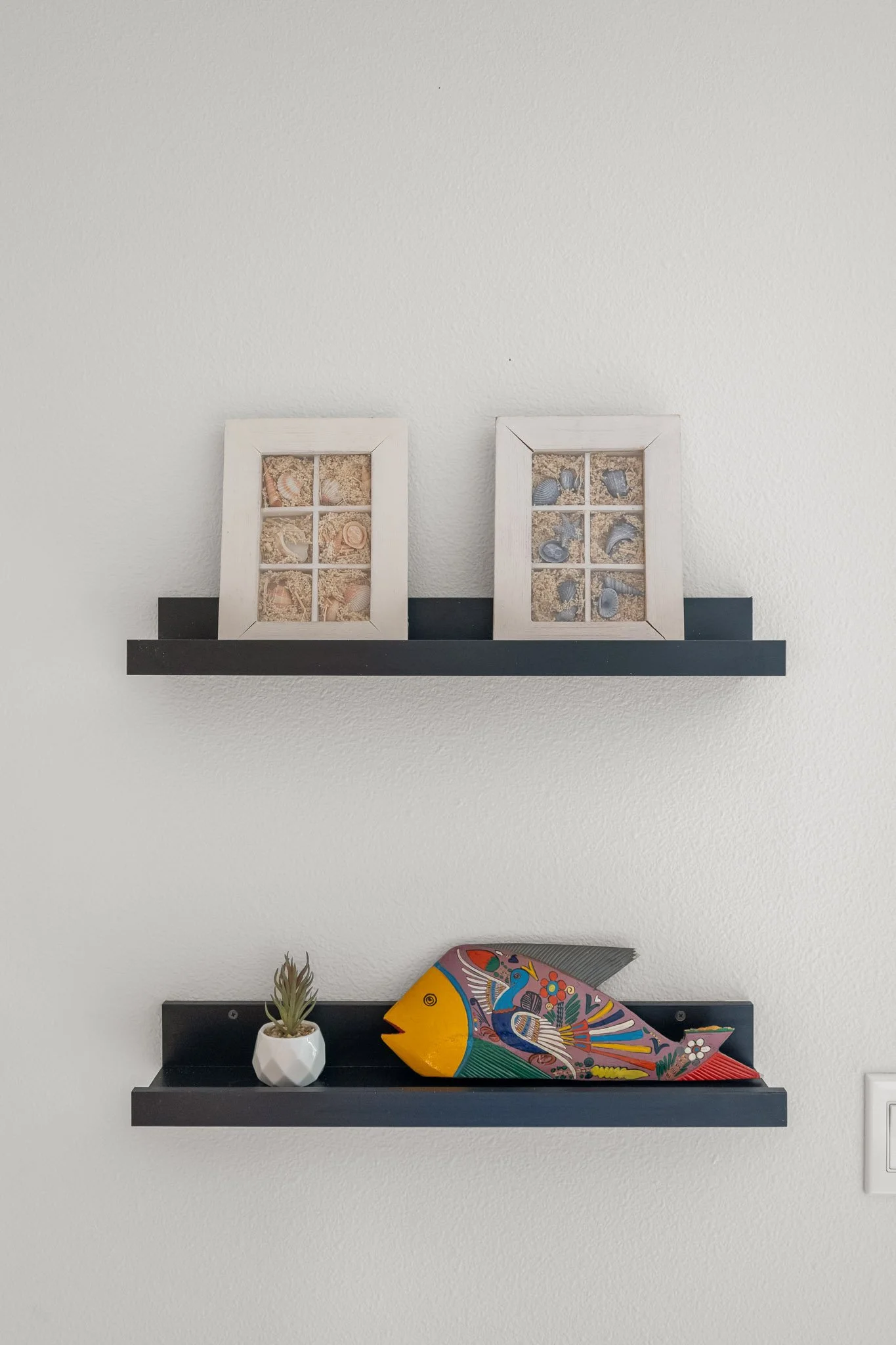 Wall-mounted black fireplace mantels with decorative items. The top shelf holds two white framed display cases with seashells and sand. The lower shelf has a small white geometric vase with a succulent and a colorful whale sculpture with intricate pa