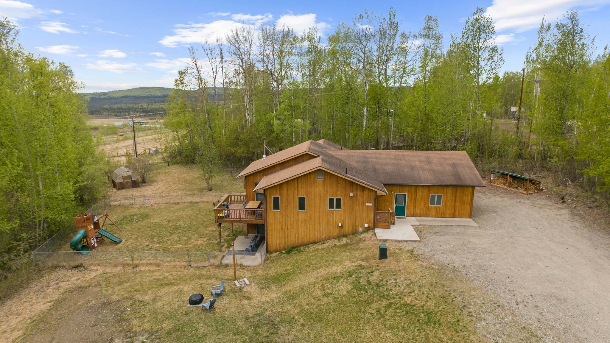 A house with a wooden exterior and a deck, surrounded by a yard with children’s playground equipment, set in a rural area with trees and distant hills under a partly cloudy sky.
